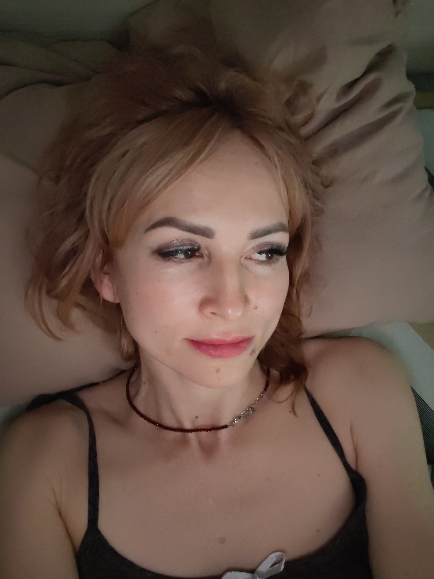 The image displays a woman lying down indoors, resting her head on a light-colored, possibly beige, pillow. She is looking to her right (viewer's left) with a subtle, contemplative expression. Her hair is light brown or blonde, appearing softly styled and slightly disheveled.
She wears makeup, including dark eyeliner, glittery eyeshadow, and a pink or berry-toned lip gloss or lipstick. Her eyebrows are neatly shaped, and she has visible long eyelashes. Several small moles are present on her face and neck. Around her neck, she wears a choker-style necklace made of small dark red beads, with a segment of silver-colored, possibly decorative, beads at the front. She is dressed in a dark-colored, thin-strapped top, likely a camisole, which features a small white bow detail near the bottom of the frame.
The lighting is soft and warm, suggesting an indoor setting, possibly in the evening or at night. A gentle light illuminates her face from the left side, highlighting her features. The scene conveys a sense of quiet relaxation or introspection. There is no visible text in the image.