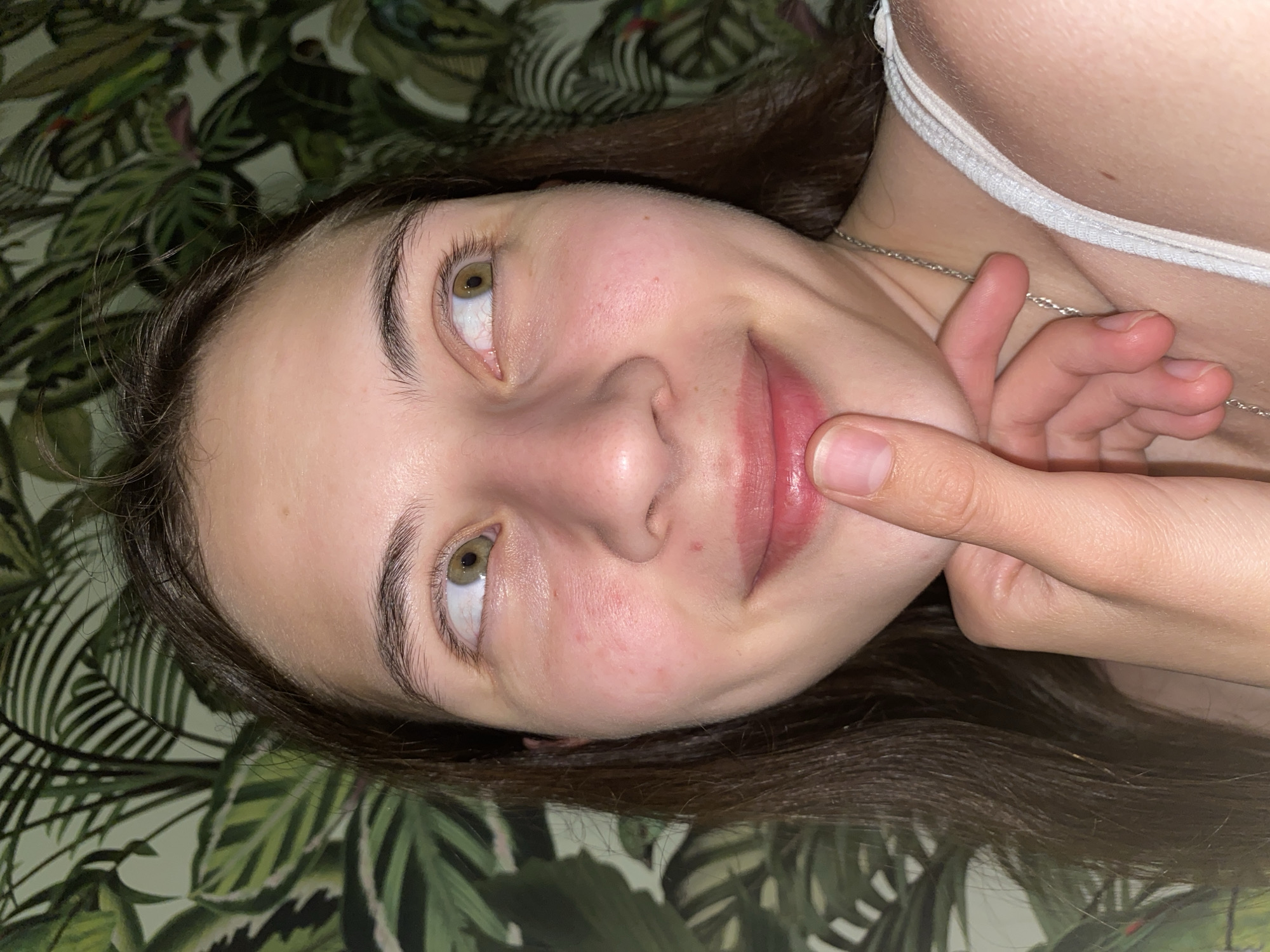 The image is a close-up portrait of a young woman with long, dark brown hair, fair skin, and hazel eyes. She is looking upwards and to her right, with a slight smile on her lips. Her right index finger is touching her lower lip. She is wearing a white tank top and a silver necklace. The background is a wall covered in green patterned wallpaper, depicting large palm leaves. There is no indication of the city of Votkinsk, Russia, or any other specific location within the image. The lighting suggests an indoor setting, possibly at night or in a dimly lit room.