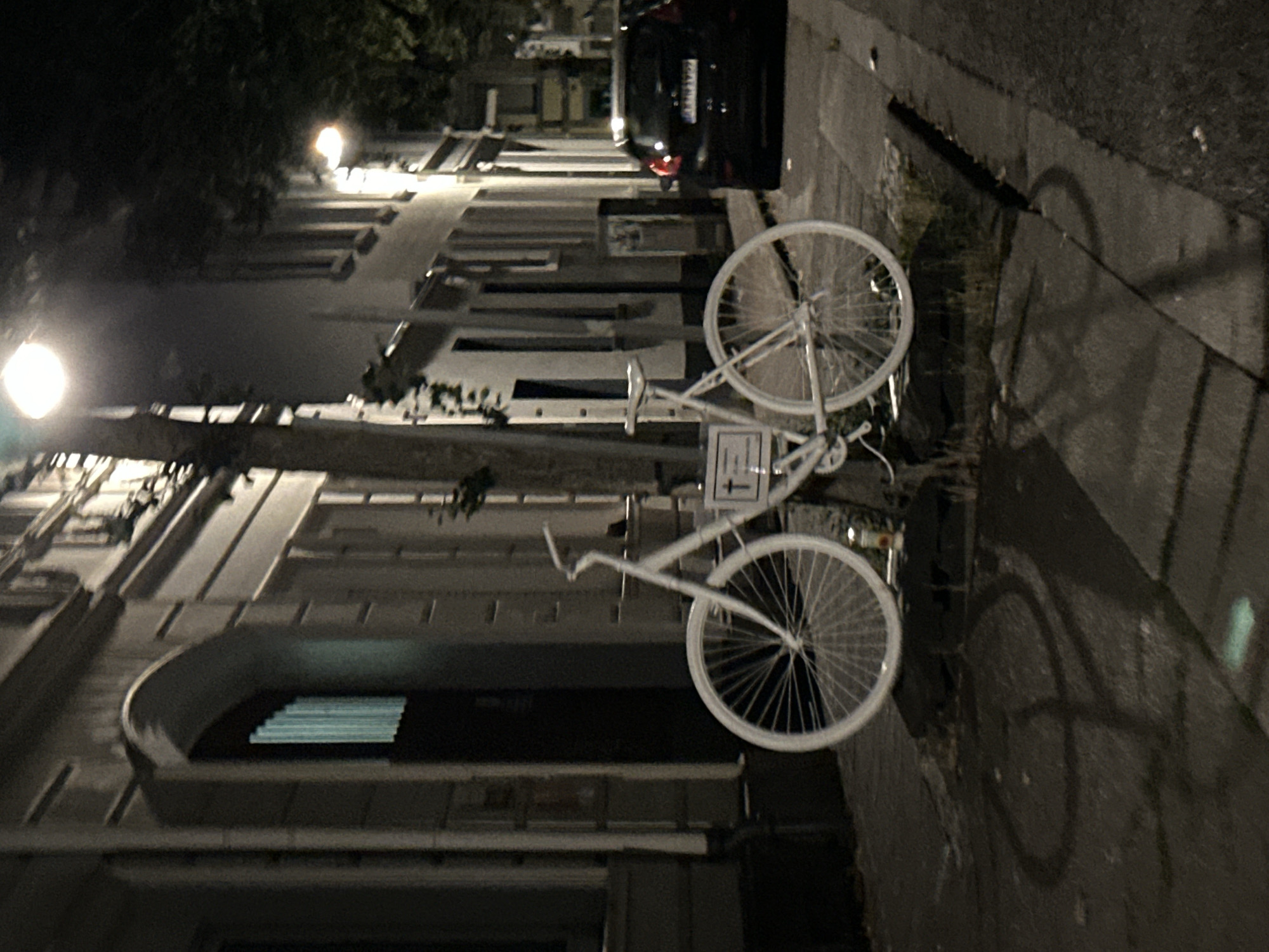 A white bicycle with a cross-shaped plaque attached to its frame is parked on a sidewalk in front of a building at night. The bicycle has a white frame, white rims, and white tires. The plaque is white with a black cross. A street light illuminates the scene. The building appears to be an old European-style structure with arched windows and a stone facade. A black car is parked on the street in the background. The sidewalk is paved with concrete. The overall impression is of a quiet, nighttime street scene in a European city. The location of the image is the Bockumer Platz in Krefeld, Germany.