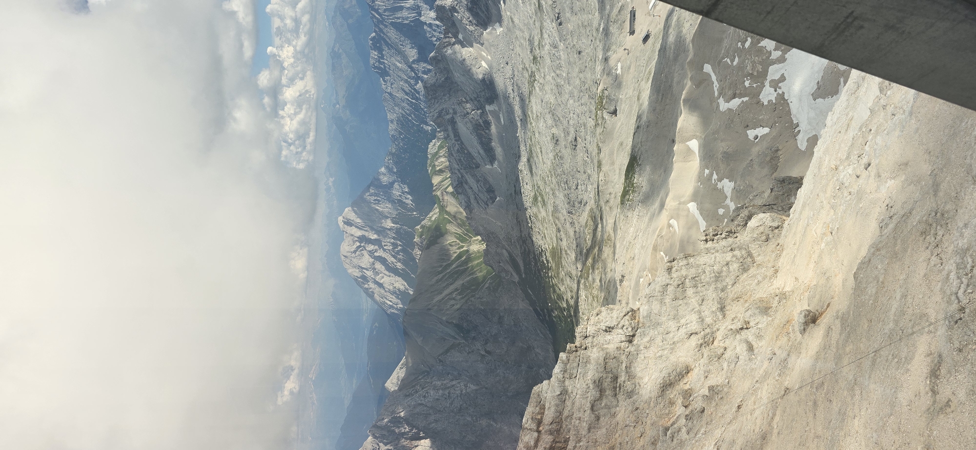 This is a photograph taken from a high vantage point, looking out over a vast mountain landscape. The foreground is dominated by a rocky cliff face, with textured grey and tan rock formations. Beyond the cliff, a wide, undulating valley floor is visible, characterized by areas of sand, gravel, and sparse green vegetation. Patches of snow are scattered across the valley floor.
In the mid-ground and background, successive layers of mountain ranges recede into the distance. The mountains are rugged and appear to be composed of grey rock, with some areas showing steep inclines and sharp peaks. The furthest mountain ranges are softer in appearance, appearing blue and hazy due to atmospheric perspective.
The sky is partially filled with large, white clouds, with patches of blue sky visible between them. Sunlight is breaking through the clouds in some areas, casting shadows on the mountains.
To the right side of the image, a dark, angled structure, possibly part of a building or viewing platform, partially intrudes into the frame. On the valley floor, small structures that appear to be buildings or possibly ski lifts are visible in the distance.
While the image depicts a mountain landscape, the specific location within Grainau, Germany, cannot be definitively determined solely from the visual information present. However, given the grandeur of the mountains, it is likely a view from a significant elevation such as the Zugspitze, Germany's highest peak, which is located near Grainau. There are no people or animals clearly visible in the photograph. The scene is one of natural, rugged mountainous terrain under a partly cloudy sky.