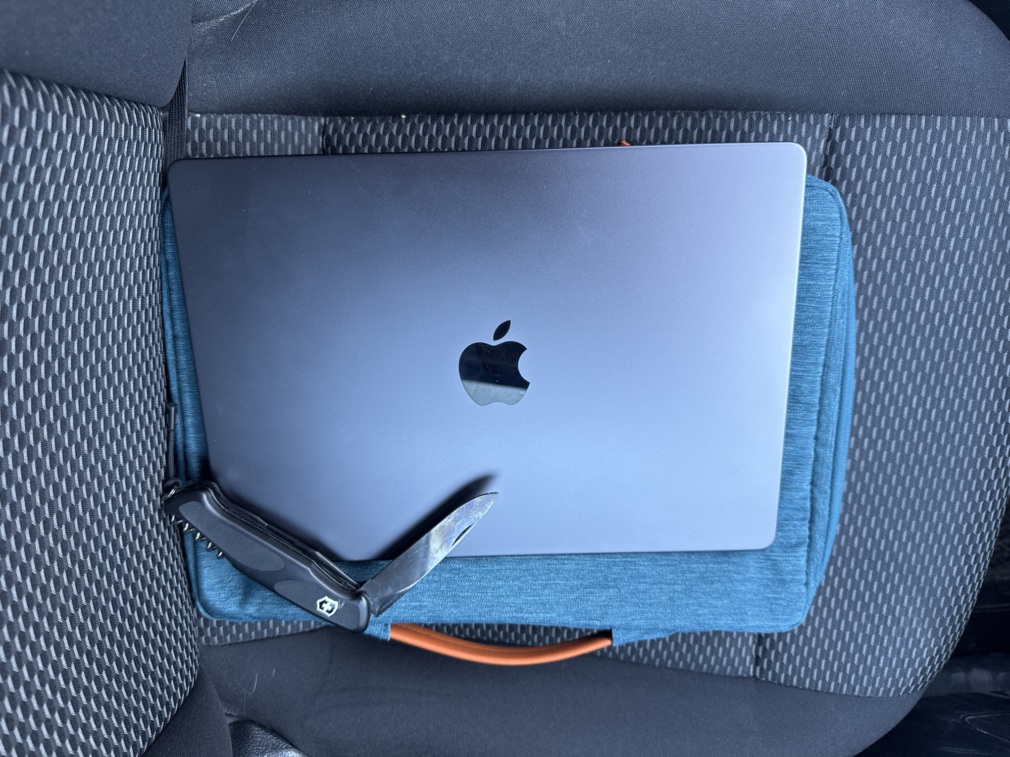 The image displays a flat lay composition of a laptop, a multi-tool knife, and a laptop sleeve resting on a car seat, likely within a vehicle in Chaikovsky, Russia.
The primary subject is a closed, dark grey (possibly Space Gray) MacBook Pro laptop, with its iconic Apple logo prominently centered on the lid. Beneath the laptop is a teal or blue fabric laptop sleeve or cushion, which features a distinctive orange-brown handle visible at the bottom center.
To the left of the laptop, a black Victorinox multi-tool knife is positioned. One of its blades is extended and appears slightly discolored or tarnished. The classic Victorinox cross and shield emblem is visible on the knife's handle.
The objects are placed on a car seat, identifiable by its dark grey/black fabric upholstery with a repeating, textured geometric pattern. The texture is visible on the seat's backrest and base. Some light dust or pet hair can be observed on the car seat fabric in various areas. The lighting is diffused and even, suggesting either an overcast day or indoor lighting conditions. No specific activity is taking place other than the static arrangement of these personal items, possibly indicating they are being transported or temporarily stored. There is no visible text other than the Apple logo and the Victorinox emblem.