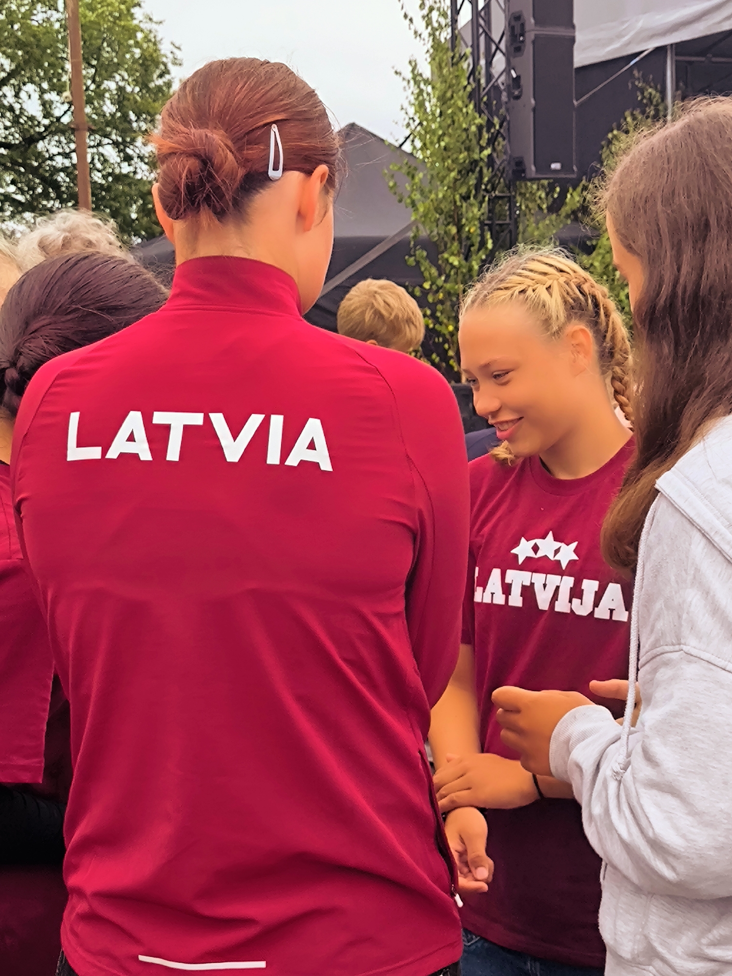 The image shows three young people, two girls and one boy, standing outdoors. The girl in the foreground is wearing a red long-sleeved shirt with "LATVIA" printed in white on the back. She has her hair in a bun secured with a silver hair clip. Another girl to her right is wearing a maroon t-shirt with "LATVIJA" and three white stars printed on it. She has braided blonde hair and is smiling and looking down. A third person, whose gender is not clearly identifiable due to their back being to the camera, is wearing a light grey hooded sweatshirt. The background includes trees, a stage with audio equipment, and a cloudy sky. The scene appears to be an outdoor event or gathering. The image was taken in Salacgrīva, Latvia.