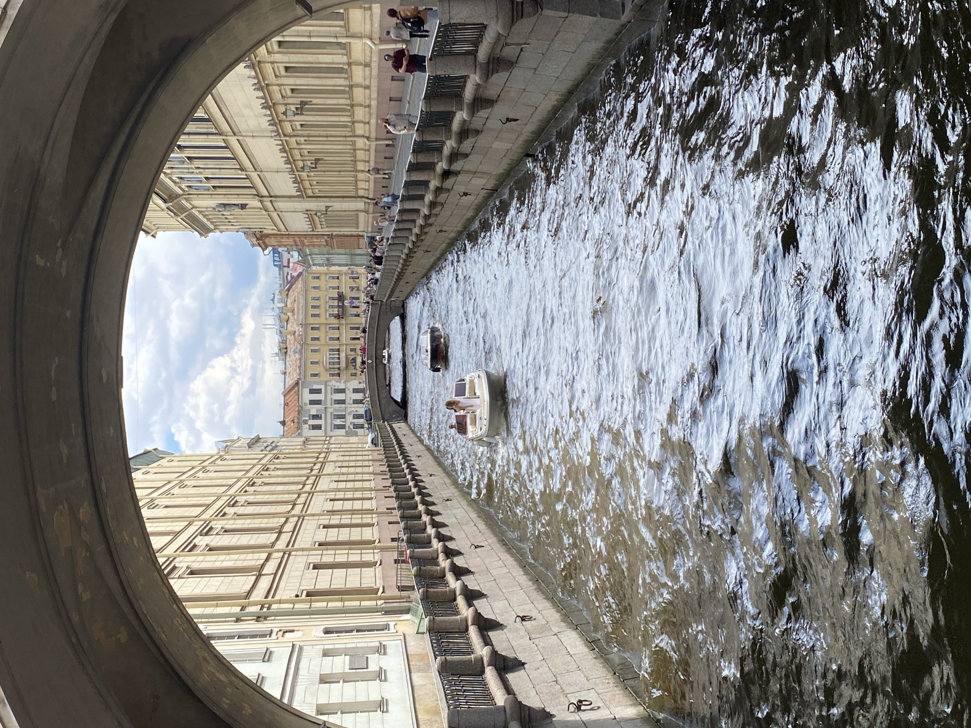 An outdoor, eye-level, vertical, medium shot captures a canal in Saint Petersburg, Russia, viewed from underneath a stone archway. The canal is lined with stone walls and ornate metal railings. On the water, a small white boat with two people aboard, one standing and wearing a white dress, moves away from the viewer. Further down the canal, another small dark boat is visible. The canal leads to a bridge in the distance, beyond which are multi-story buildings with distinct architectural styles, some featuring red roofs. Pedestrians are seen walking along the canal's edge and on the bridge. The sky above is partly cloudy. The scene depicts a sunny day in the city.