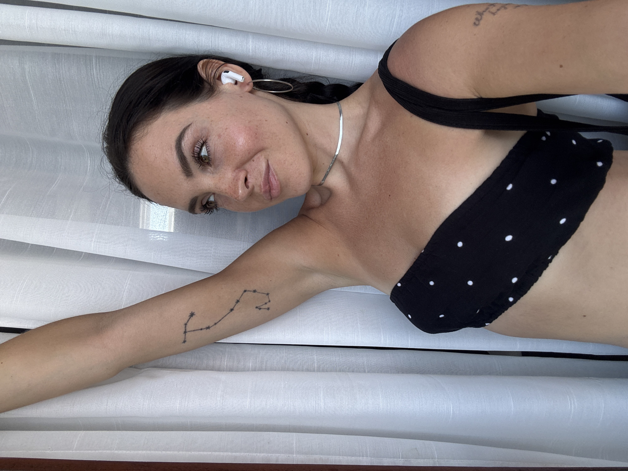 A woman is photographed in a portrait-style indoor setting. She is looking off to the right, with her left arm raised and resting against a white curtain. Her hair is dark and braided down her back. She is wearing a black polka-dot strapless top and a thin silver necklace. She has a constellation tattoo on her upper left arm and another faint tattoo on her right arm. She is wearing white wireless earbuds and large hoop earrings. The background consists of white curtains. The image is sharp and well-lit, with a shallow depth of field.
