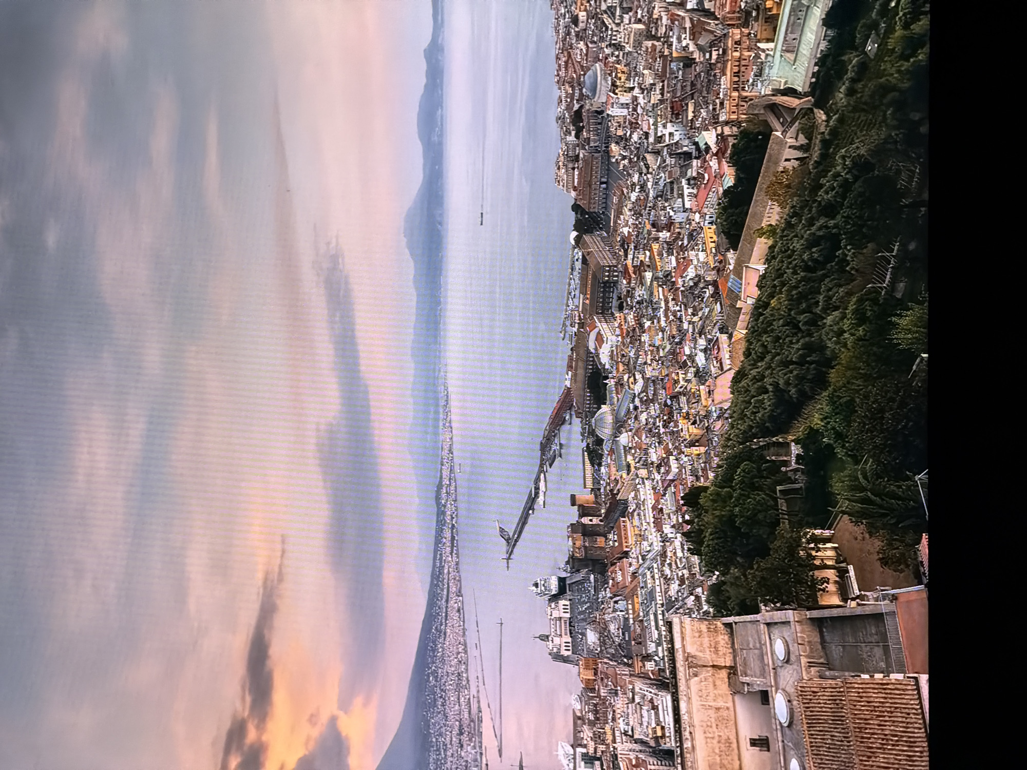 The image is a panoramic aerial view of a coastal city with a large body of water and a mountain range in the background. The sky is painted with soft hues of pink, orange, and blue, indicating either sunrise or sunset. The city itself is densely populated with buildings featuring red-tiled roofs, common in Mediterranean architecture.
In the foreground, a large building with a terra cotta tiled roof is visible. This building has a courtyard with a fence and appears to be situated on a hillside overlooking the city and the water. Further down the hillside, lush green trees and vegetation are abundant, with some walking paths and what appear to be sculptures or statues.
The city stretches out towards the water, which has a prominent pier or breakwater extending into it. Several boats, including a large cruise ship, are docked or navigating the waters. In the distance, a mountain range forms the horizon, with the silhouette of Mount Vesuvius clearly identifiable as the prominent peak on the left.
The provided location information states that the image is of the city of Chirchik, Uzbekistan. However, based on the visual evidence, particularly the presence of Mount Vesuvius and the architectural style of the city, the scene is clearly not Chirchik, Uzbekistan. The image is instead a view of Naples, Italy, with Mount Vesuvius in the background.