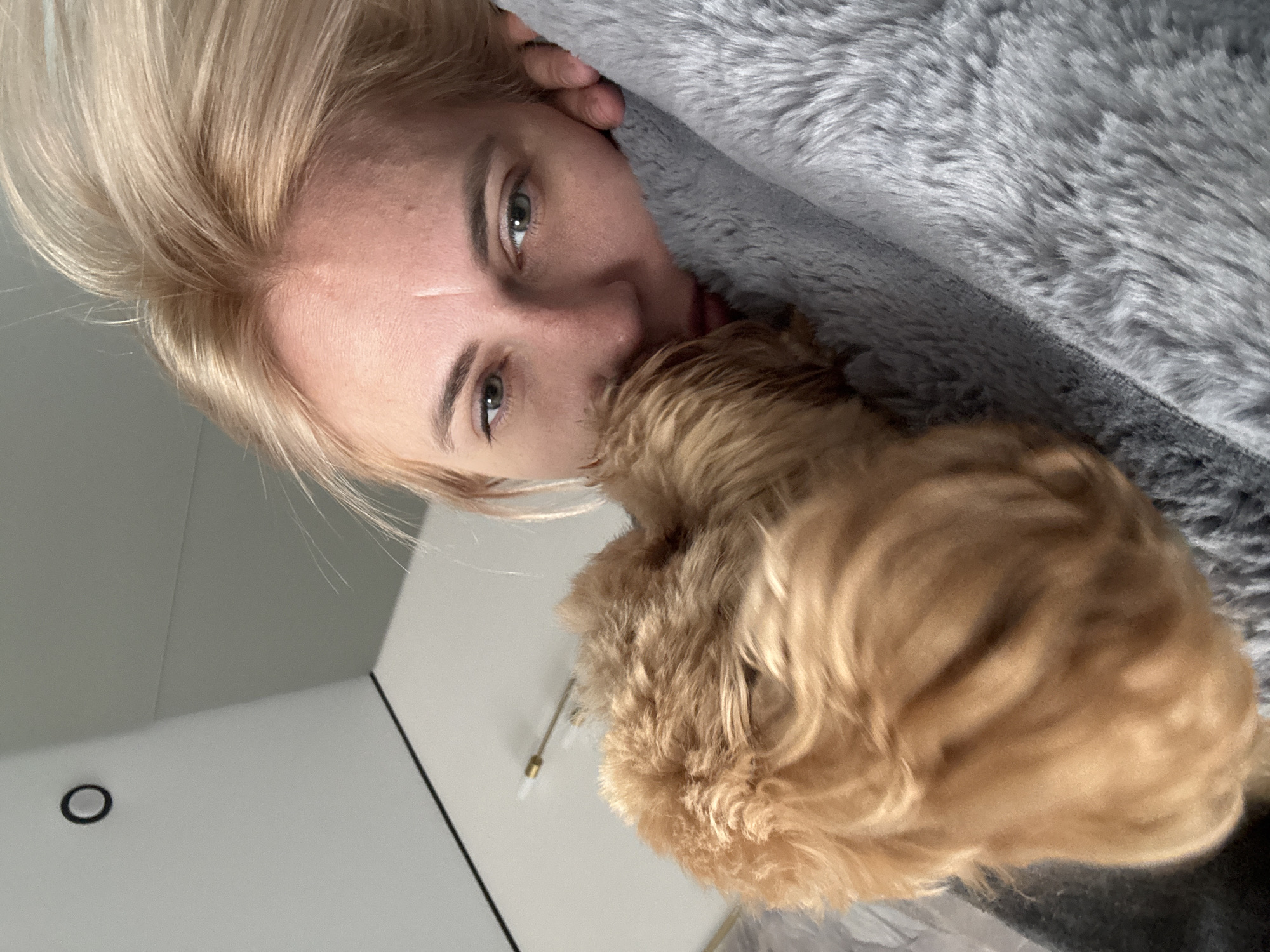 A woman with short blonde hair and green eyes is lying down next to a fluffy, light-brown, small dog. She is partially covered by a soft, gray blanket. The scene also shows part of a white wall with a black circle and vertical black line, suggesting a modern interior. The location is not discernible from the image.