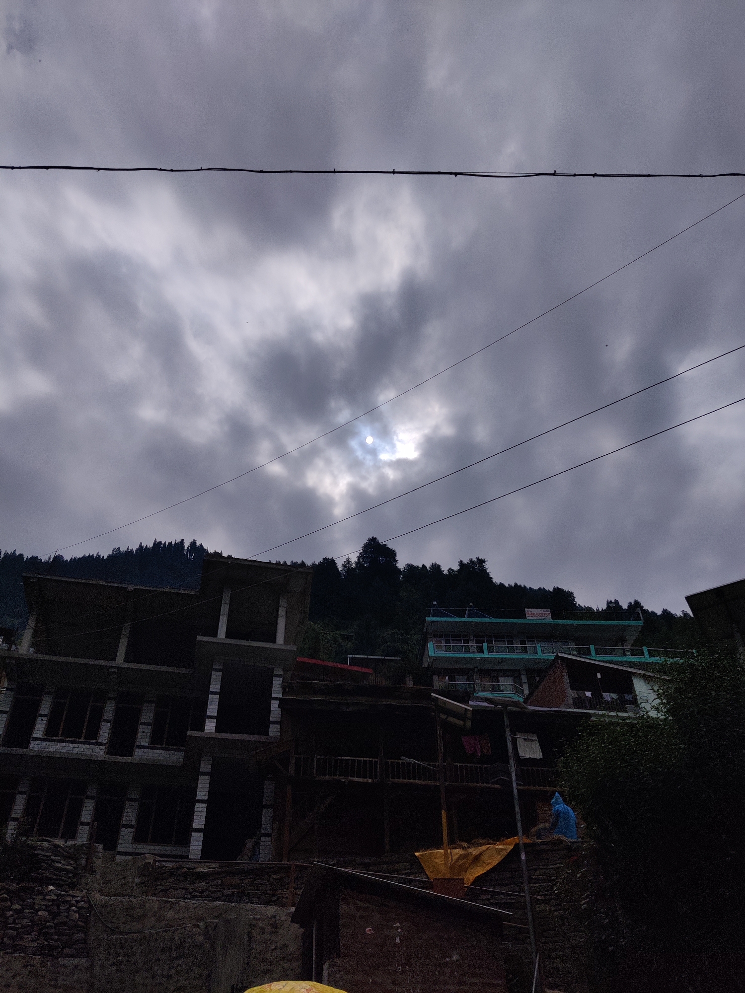 The image captures a view of Vashisht, India, with overcast skies and partial sunlight filtering through the clouds. In the foreground, a multi-story building under construction is visible, with concrete pillars and unfinished walls. To the right, another building with a turquoise roof and balconies is situated on a slope, and a person wearing a blue hooded garment is seated on a stone wall. Behind the buildings, a dense forest covers the hillside. Power lines crisscross the sky, adding to the scene's urban mountain setting. The overall atmosphere is subdued due to the cloudy weather.