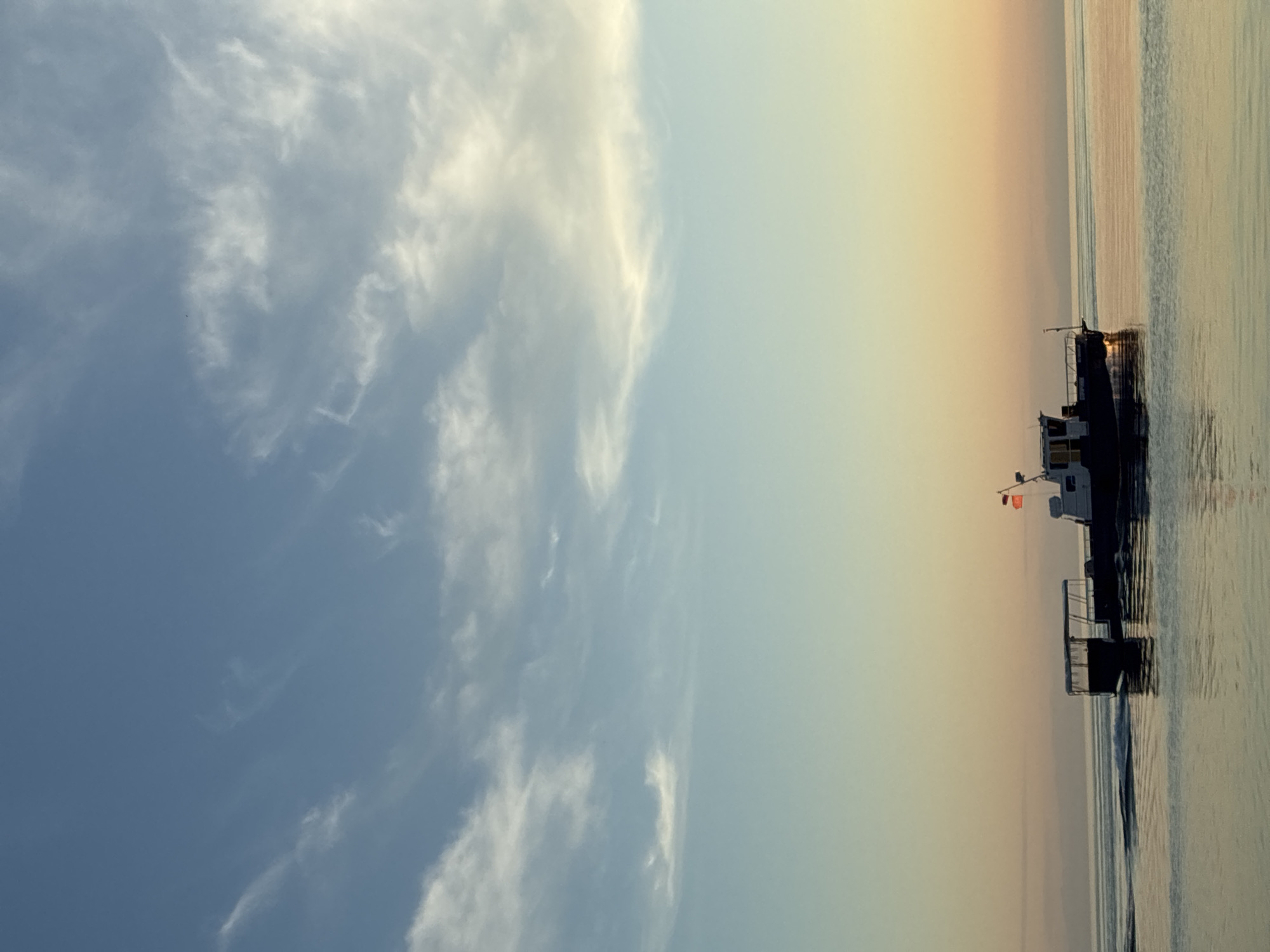 A boat sails on the water under a sky with sparse clouds. The boat has a covered seating area and a smaller cabin at the rear. The water is calm, with a gentle wake trailing behind the boat. The sky transitions from a pale blue at the top to a warm orange and pink hue near the horizon, suggesting either sunrise or sunset. In the distance, a faint outline of land or mountains is visible. The scene is serene and likely depicts Lake Baikal near Listvyanka, Russia, given the typical landscape and sky conditions. There are no people visible in the image.