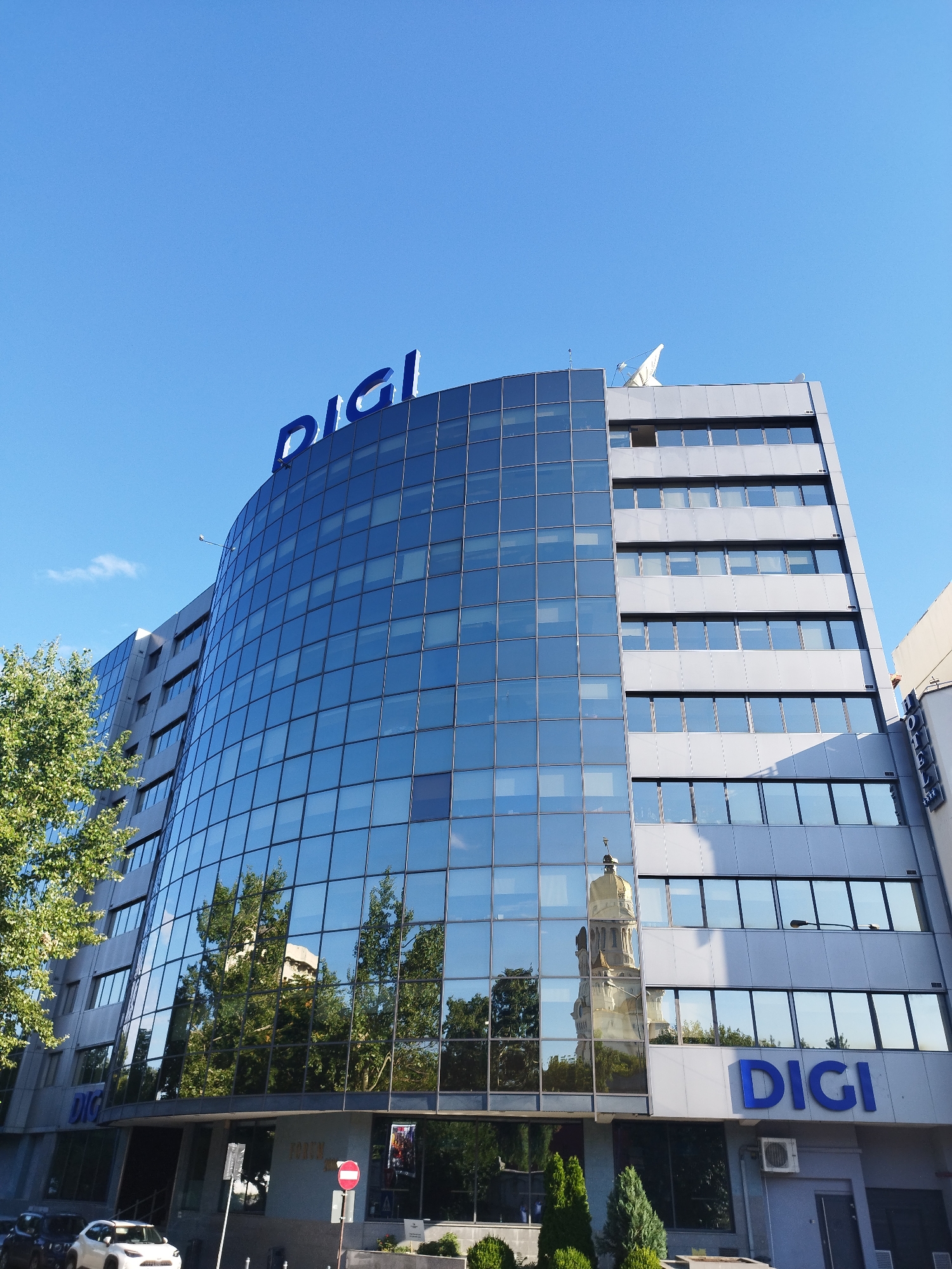 The image shows the DIGI headquarters building in Bucharest, Romania, on a clear, sunny day. The modern building features a curved facade of blue-tinted glass windows, reflecting the surrounding trees and a church tower. The DIGI logo is prominently displayed in large blue letters on the top of the building and again on the lower section. To the right of the DIGI building, part of a hotel is visible, with the word "HOTEL" written vertically. The street level shows cars parked, a street lamp, and some greenery. The sky is a vibrant blue, with a few wispy clouds.