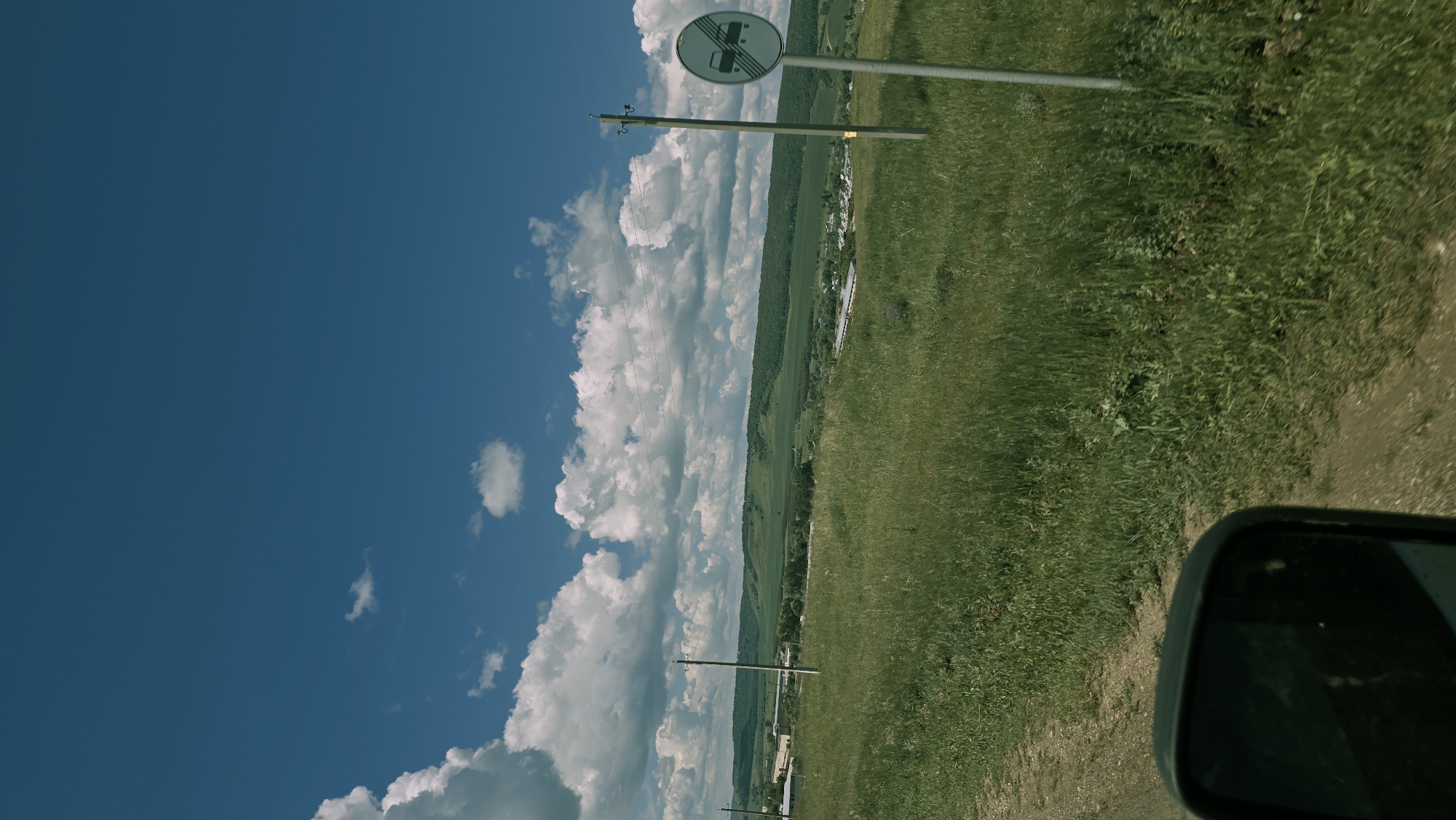 The image depicts a rural landscape under a bright blue sky with scattered white clouds. The foreground shows a dirt road and a side-view mirror of a vehicle. A grassy field extends into the middle ground, dotted with tall grass and wildflowers. Two utility poles are visible in the field, one with a circular sign indicating "End of Road" or a similar traffic restriction. In the distance, rolling green hills are covered with forests, and a small village is nestled in the valley. The scene appears to be located in the village of Kudashevo, Russia, judging by the provided context, characterized by its natural beauty and a sense of peaceful rural life. There are no people visible in the image. The lighting suggests it is daytime, likely late morning or early afternoon.