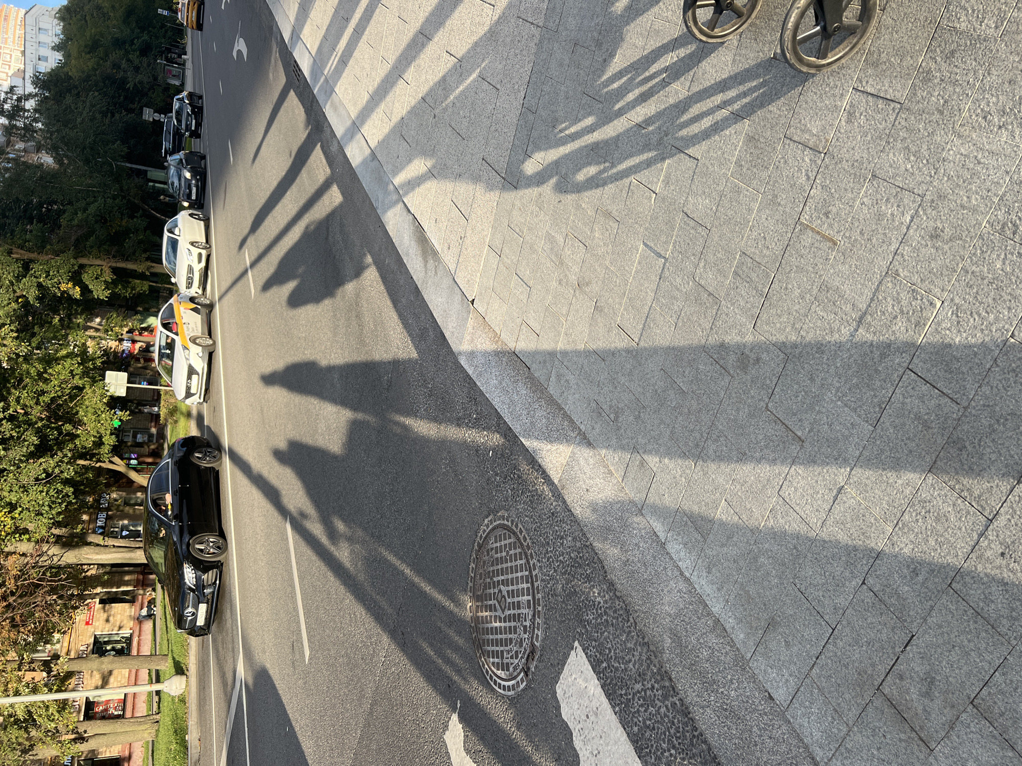 A black Mercedes sedan is seen driving on a street in Moscow, Russia. The car is in the left lane of a multi-lane road. In the background, there are trees and buildings. The shadows of people are cast on the road and sidewalk. The sidewalk is paved with granite tiles. Two wheels of what appears to be a stroller are visible in the lower right corner of the image.