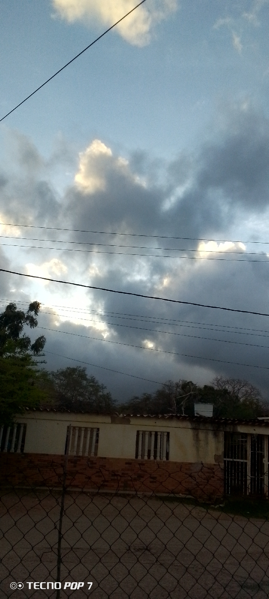 The image depicts a cloudy sky above a low-rise building, with power lines traversing the frame. The building appears to be a residential or commercial structure with barred windows and a tiled roof. A chain-link fence is visible in the foreground. The scene is likely set in Altagracia, Venezuela, as indicated by the "TECNO POP 7" watermark, which is a mobile phone brand, and the style of the building and surrounding vegetation. The lighting suggests it is either early morning or late afternoon, with the sun partially obscured by the clouds. No people are visible in the image. The overall impression is one of a quiet, possibly somewhat overcast day in a residential area.