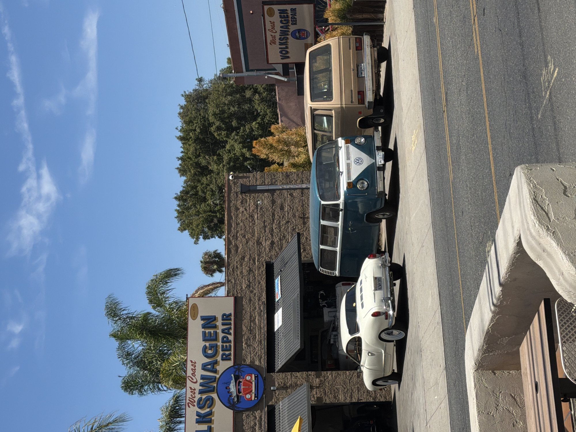 The image captures an exterior view of "West Coast Volkswagen Repair" in Long Beach, California, on a sunny day. The repair shop features a brick facade with a large overhead door open to reveal the interior. Parked in front of the shop are three classic Volkswagen vehicles: a white Karmann Ghia convertible on the left, a teal and white Volkswagen bus in the middle, and a tan Volkswagen van on the right. The sky is bright blue with a few wispy clouds. Palm trees are visible in the background. A portion of an outdoor seating area with a table and chairs is in the foreground, suggesting the establishment may also offer a waiting area for customers. The overall scene depicts a well-maintained auto repair shop specializing in vintage Volkswagen vehicles.