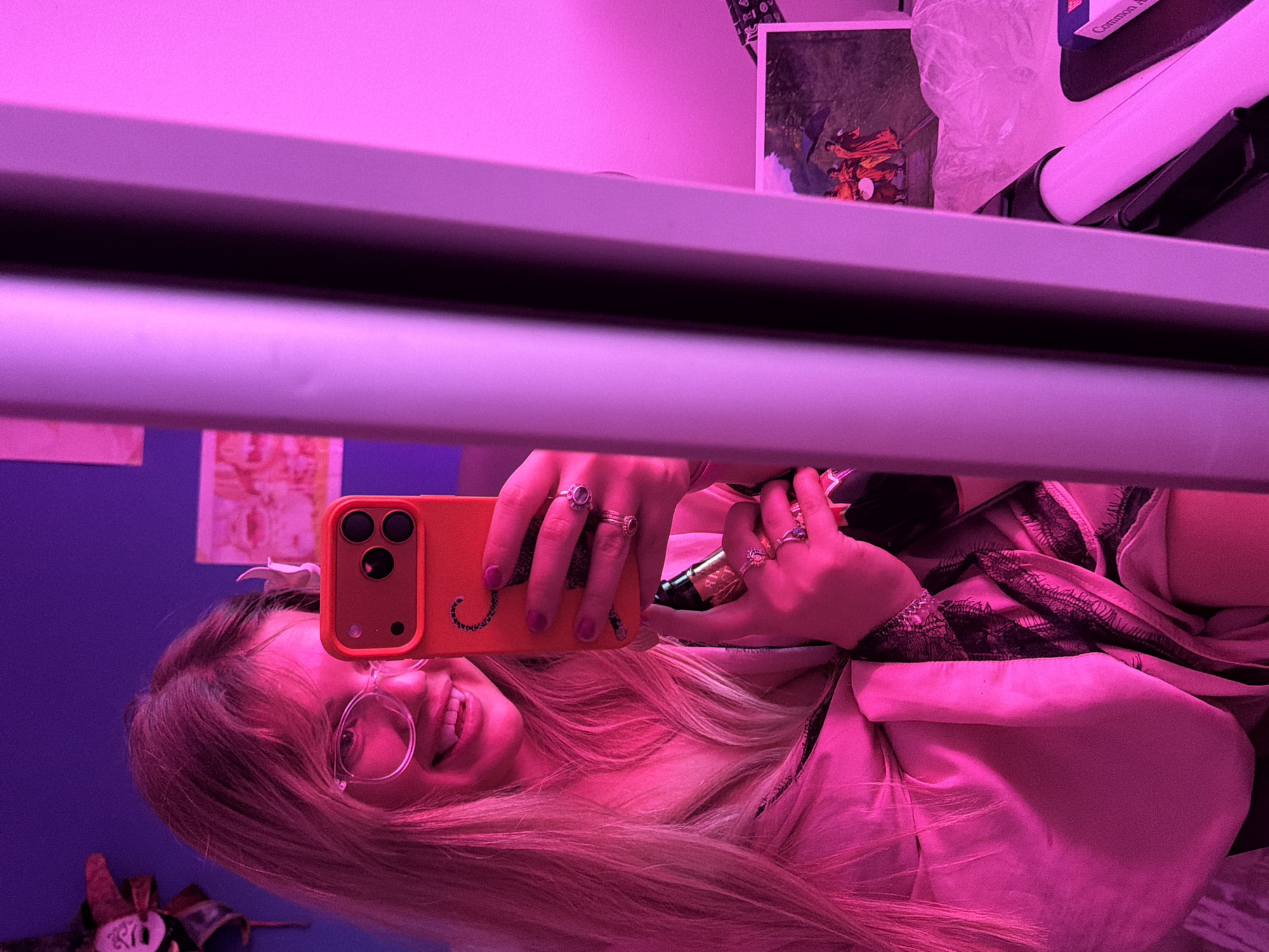 A woman is taking a selfie in a room with pink lighting. She is holding an orange smartphone and smiling. The background includes a desk with items such as a picture frame, a tissue box, and other objects. The location is specified as being within the city of Большая Шильна, Russia.