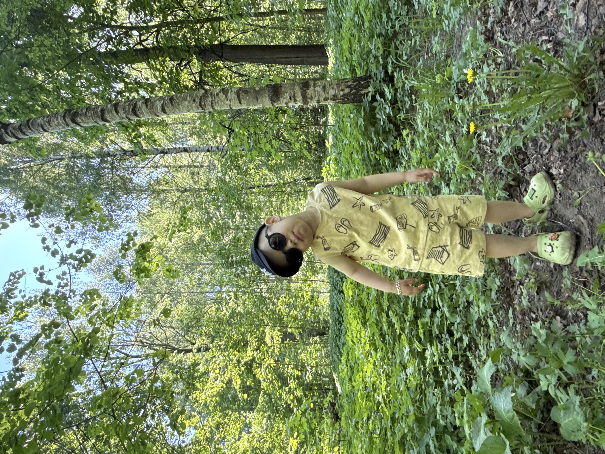 A child wearing a yellow outfit, a black hat, and sunglasses stands in a forest area. The child's outfit has various motifs, including seashells and palm trees. The child is also wearing green Crocs-style shoes. They have a bracelet on their left wrist. The forest is densely vegetated with green plants and a few yellow flowers. A birch tree with distinctive white bark is visible in the background. The sky is visible through the trees in the background. The location appears to be in a wooded area, and the photo's orientation may be rotated. It is not possible to confirm the location as Rusanovo, Russia based on visual elements alone.