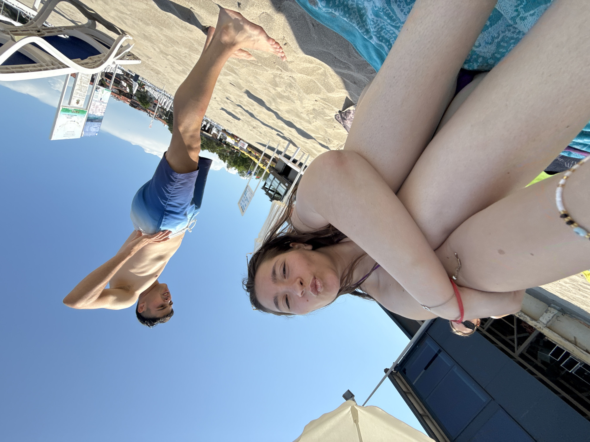 The image, rotated counter-clockwise by 90 degrees, captures a bright, sunny day at a beach in Sozopol, Bulgaria. In the immediate foreground, a young woman is seen from the chest up, her face inverted relative to the ground. She looks towards the camera with a playful "duck face" expression, her long brown hair flowing around her shoulders. She wears a purple bikini top, and her left arm, adorned with a red bracelet, is bent, resting near her chest. Her bare legs are visible in the upper right quadrant, resting on a patterned blue and white towel spread on the sand.
Further back and to the left, a young man is also seen inverted against a clear blue sky, wearing blue and white swimming shorts. He is shirtless, with his hands on his hips, looking towards the camera with a relaxed expression. The ground is sandy, with distinct shadows indicating strong daylight, suggesting it is midday or early afternoon. Several white plastic sun loungers are visible on the sand, along with what appear to be information boards or signs with illegible text. In the background, various beach structures, including a building resembling a beach bar or cafe, and a white umbrella, are present. The scene depicts a leisurely summer day at the seaside.