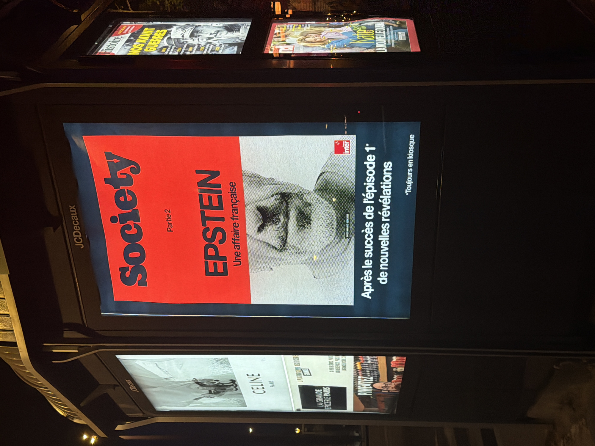 The image captures a street-level advertising display in Paris, France, under nighttime conditions. The main focus is on a large poster advertising "Society Partie 2," which features the name "EPSTEIN: Une affaire française" prominently displayed in red over a grayscale image of a man. To the right of the poster, French text states, "Après le succès de l'épisode 1* de nouvelles révélations *Toujours en kiosque" (After the success of episode 1* new revelations *Always in newsstands).
Above this poster, other advertisements are visible, including magazine covers: "Histoire NOS'AVANT GUERRES" and "Kate LA NOUVELLE DIANA." Below the Epstein poster, another row of ads includes one for "CELINE PARIS" and "LA GRANDE EPICERIE PARIS." The name "JCDecaux" is visible on the left side of the display unit, indicating the advertising company managing the space. The overall environment appears to be an urban setting at night.