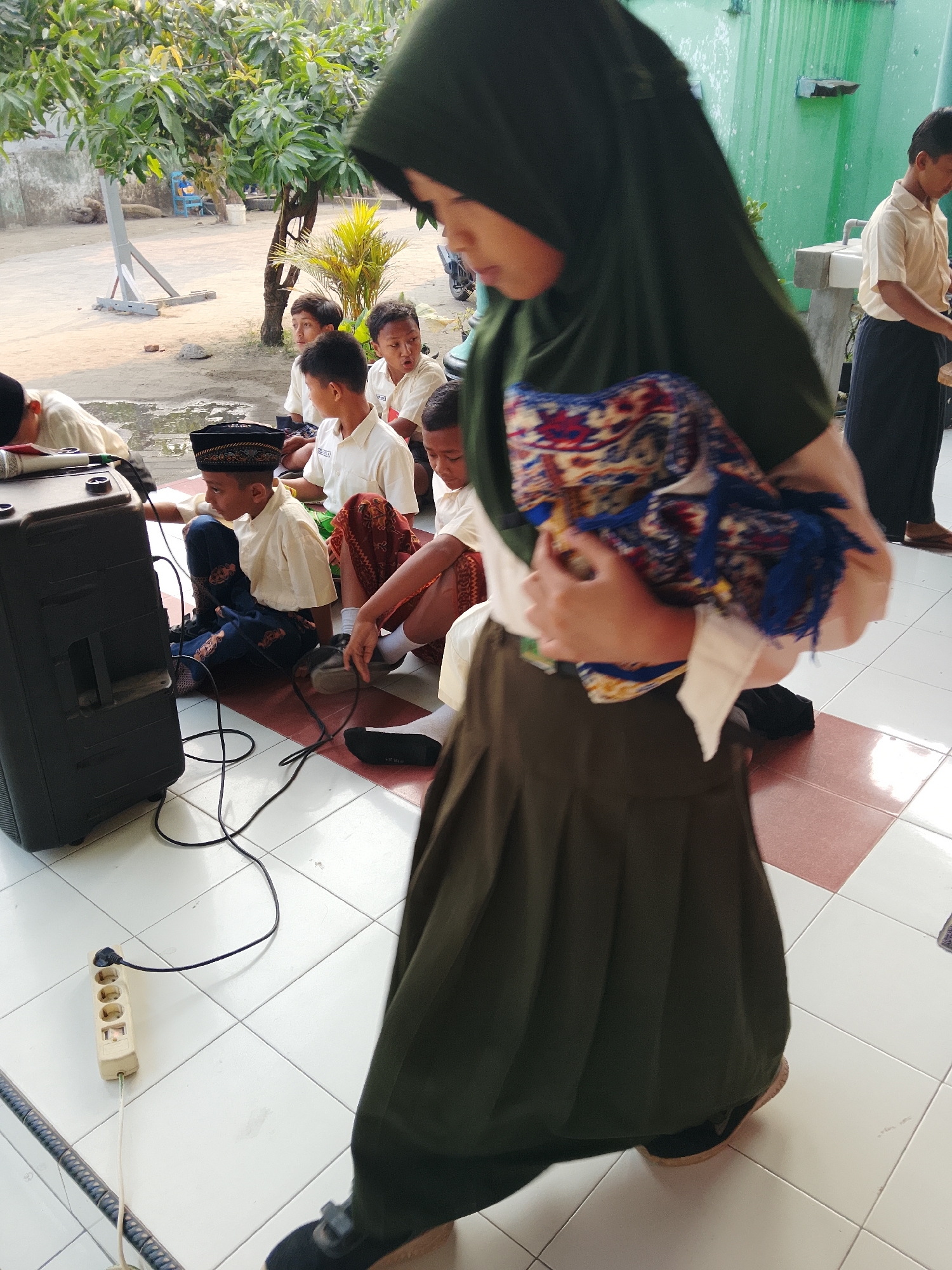 In Kepuh, Indonesia, a group of young boys, some wearing peci hats and white shirts with patterned pants or sarongs, sit on a tiled floor. In the foreground, a young person in a dark green hijab and dark skirt walks away from the camera, holding a patterned textile. To the left, a large black speaker is connected to a power strip via a black cord, with another white cord extending from the strip. The background shows a courtyard with trees, a green wall, and a sink area.