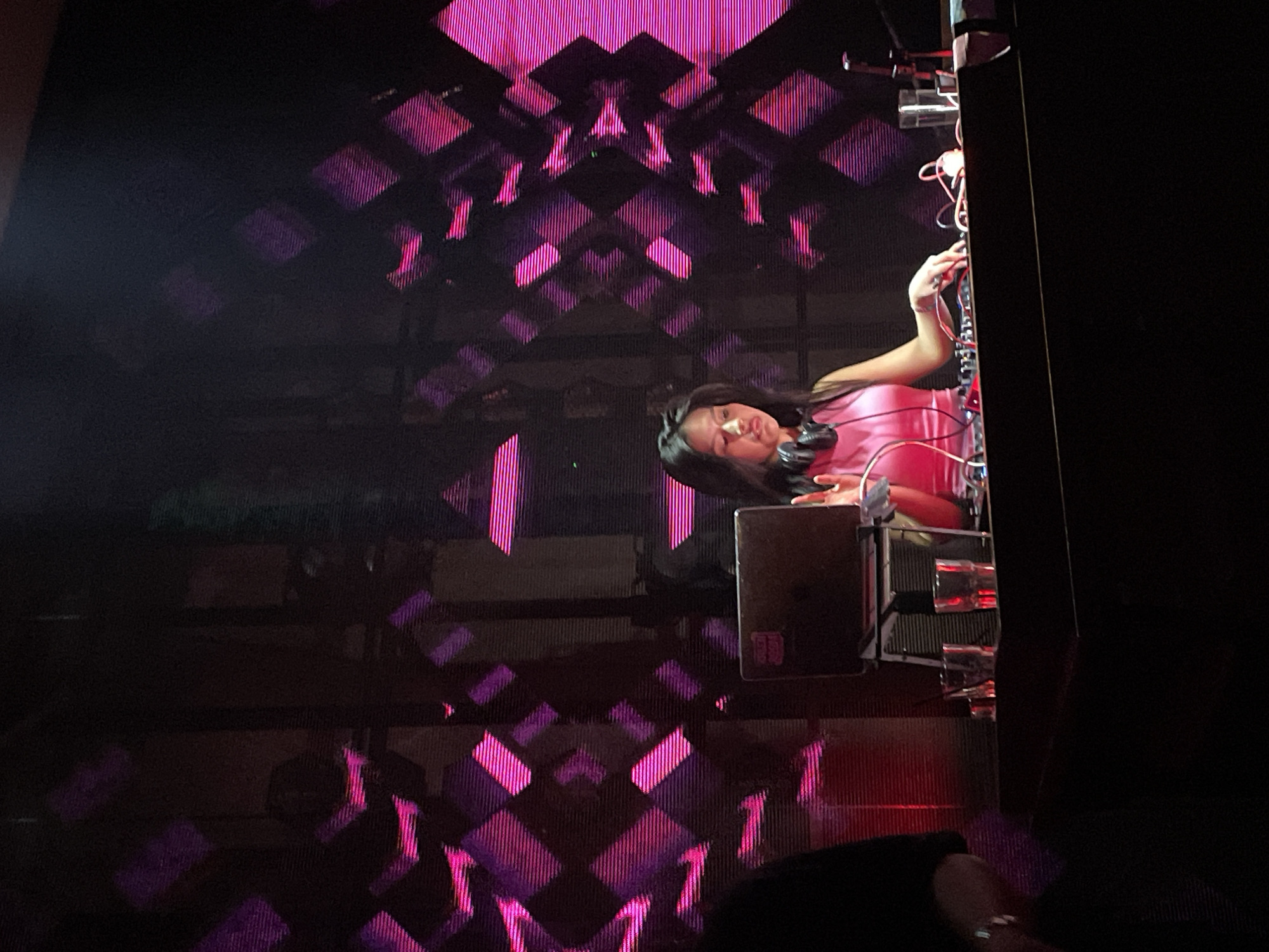 In Central Jakarta, Indonesia, a female DJ, wearing headphones and a pink sleeveless top, performs at a DJ booth. Her hands are positioned over the DJ equipment, suggesting she is actively mixing. A laptop with a sticker is visible on the left side of the booth, along with several glasses. The background features a large screen displaying a geometric pattern of pink and purple lights, creating a vibrant and energetic atmosphere. The scene is dimly lit, with spotlights highlighting the DJ and her equipment.