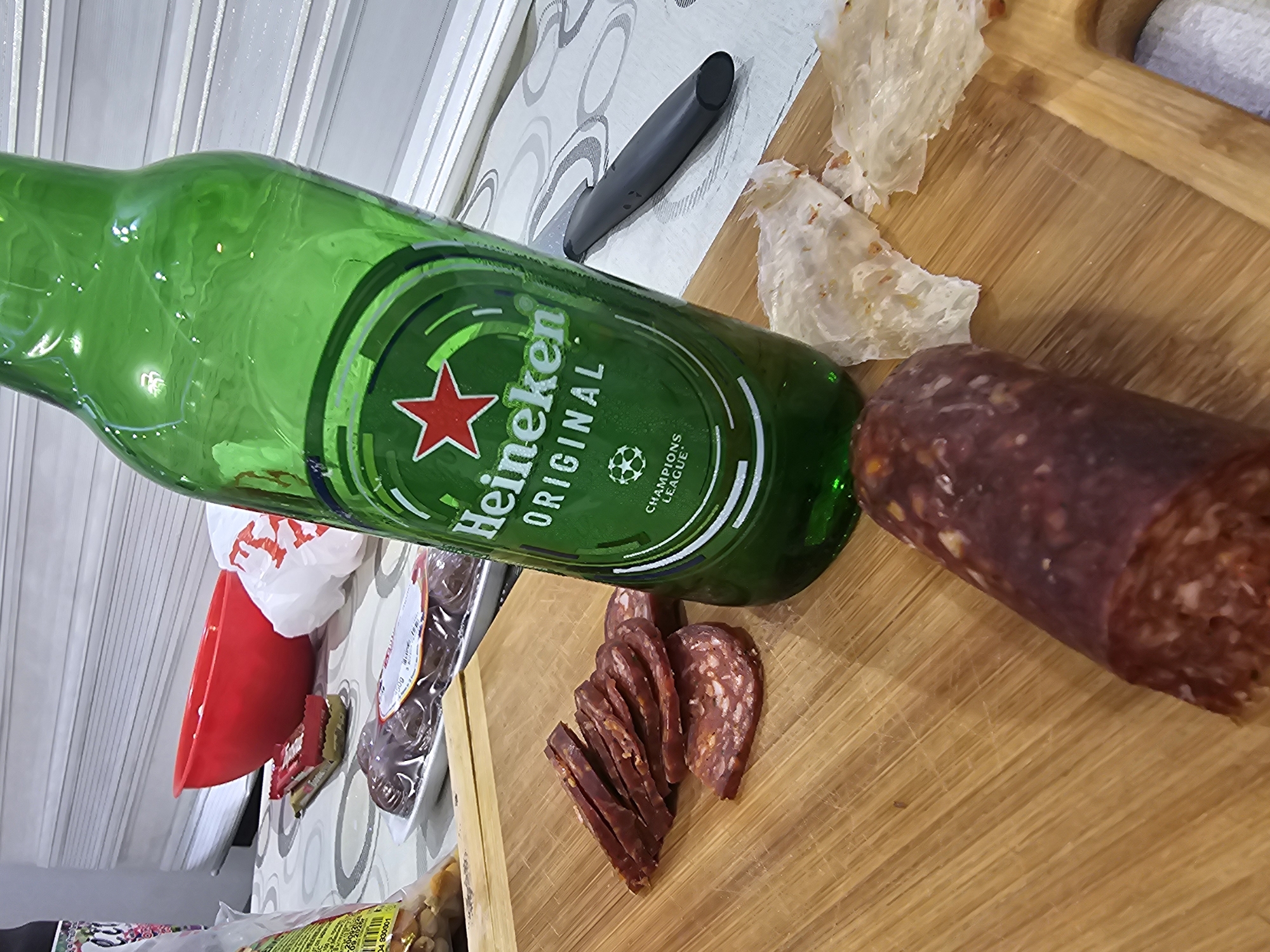 An overhead, heavily tilted shot displays an arrangement of food and drink on a light wooden cutting board and a patterned surface, likely a table or counter. The scene features a green glass bottle of Heineken beer prominently in the center-left. Its label is clearly visible, featuring a red star, the text "Heineken ORIGINAL," and a "CHAMPIONS LEAGUE" logo with a football icon.
To the right of the bottle, a thick, dark red-brown sausage rests on the cutting board. Several thin, round slices of the same sausage are fanned out next to it. Also on the cutting board are several irregular, flaky pieces of light-colored flatbread or crackers. A black-handled knife is partially visible on the patterned surface behind the beer bottle.
In the blurred background on the left, a red plastic bowl and a white plastic bag are discernible. The patterned surface, possibly a tablecloth or countertop, extends across the scene. Further in the background, a white surface with vertical stripes is visible. The overall impression is an informal snack or meal setup, with the items seemingly prepared or about to be consumed. The location context is Karnobat, Bulgaria.