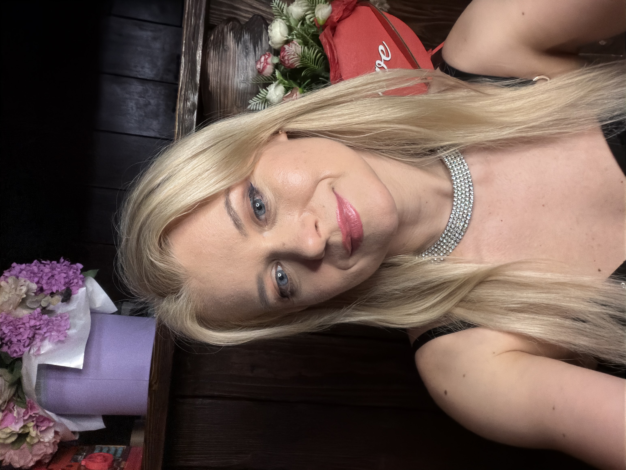 A blonde woman with blue eyes and pink lipstick is pictured in a selfie, looking at the camera with a slight smile. She has on a black top and a sparkling choker necklace. Behind her, on a wooden shelf, are two floral arrangements. The one on the left is in a lavender bucket, and the one on the right is in a red heart-shaped box with the word "love" written on it. The background appears to be a wooden wall. There is no information to suggest this location is in Sochi, Russia.