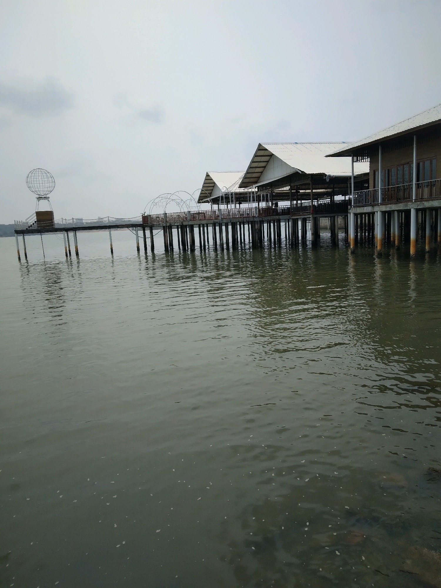 The image shows a waterfront dining or recreational area in Batam, Indonesia. The structures are built on stilts over the water, with a walkway connecting them. The buildings have a traditional Indonesian architectural style with sloped, white roofs. A prominent feature on the walkway is a globe-like sculpture. The water appears murky, with some debris visible. The sky is overcast, suggesting a cloudy day. The location is likely a tourist attraction given the unique structure and the presence of what appears to be a globe monument.