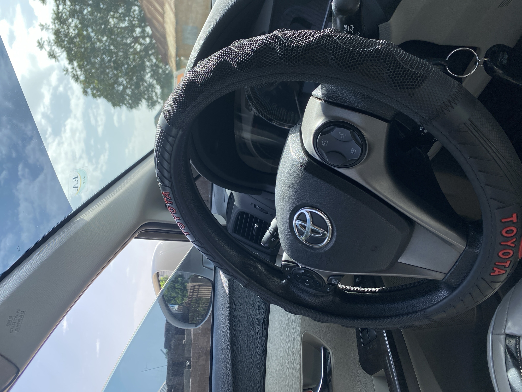 The image provides a close-up interior view of a Toyota car's steering wheel and dashboard, suggesting the car is parked. Outside the car, through the windshield and side window, a typical Nigerian semi-urban or rural setting is visible. The background shows a cluster of buildings with corrugated iron roofs, indicative of residential structures. A large tree with dense foliage is also prominent, casting shadows. The sky is bright with scattered clouds, suggesting daytime. On the steering wheel, the "TOYOTA" logo is clearly visible, along with a steering wheel cover. The car's interior appears clean and well-maintained. The presence of the dashboard, steering wheel, and door panel suggests the photo was taken from the driver's or front passenger's seat. The overall impression is of a vehicle parked in a residential area under a clear, sunny sky.