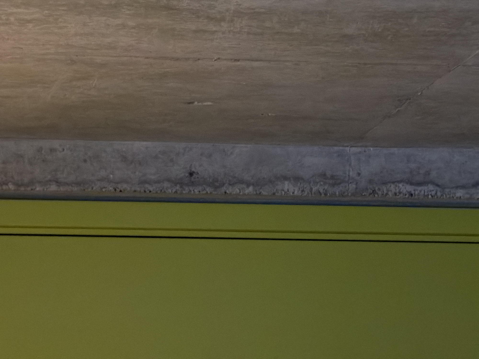 The image shows a section of a building interior in Chkalov, Armenia. A green painted wall is at the bottom of the frame. Above the wall is a horizontal concrete beam or lintel with a rough texture. Above the beam is a concrete ceiling, exhibiting cracks and imperfections. There are no people visible.
