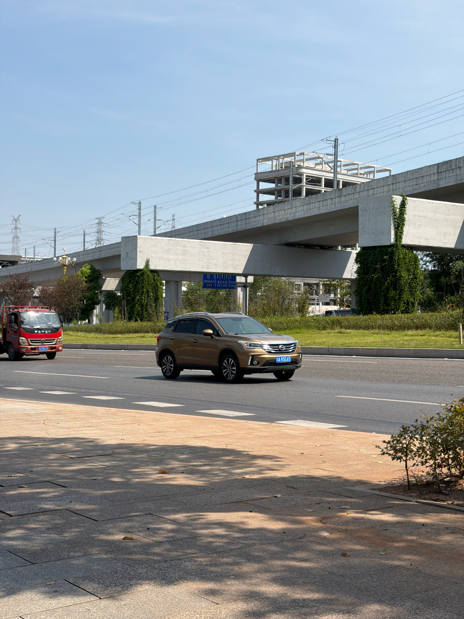 In Jiangjin, China, a gold SUV with license plate "ARSE65" drives on a multi-lane road under a bright, clear sky. To the left of the SUV, a red Foton truck is parked, also on the road. Elevated railway tracks and a partially constructed building are visible in the background, casting shadows on the road and sidewalk. Lush green vegetation, including trees and bushes, lines the sides of the road and climbs up the support pillars of the elevated structure. The sidewalk in the foreground is paved with grey tiles and illuminated by dappled sunlight filtering through overhead trees.