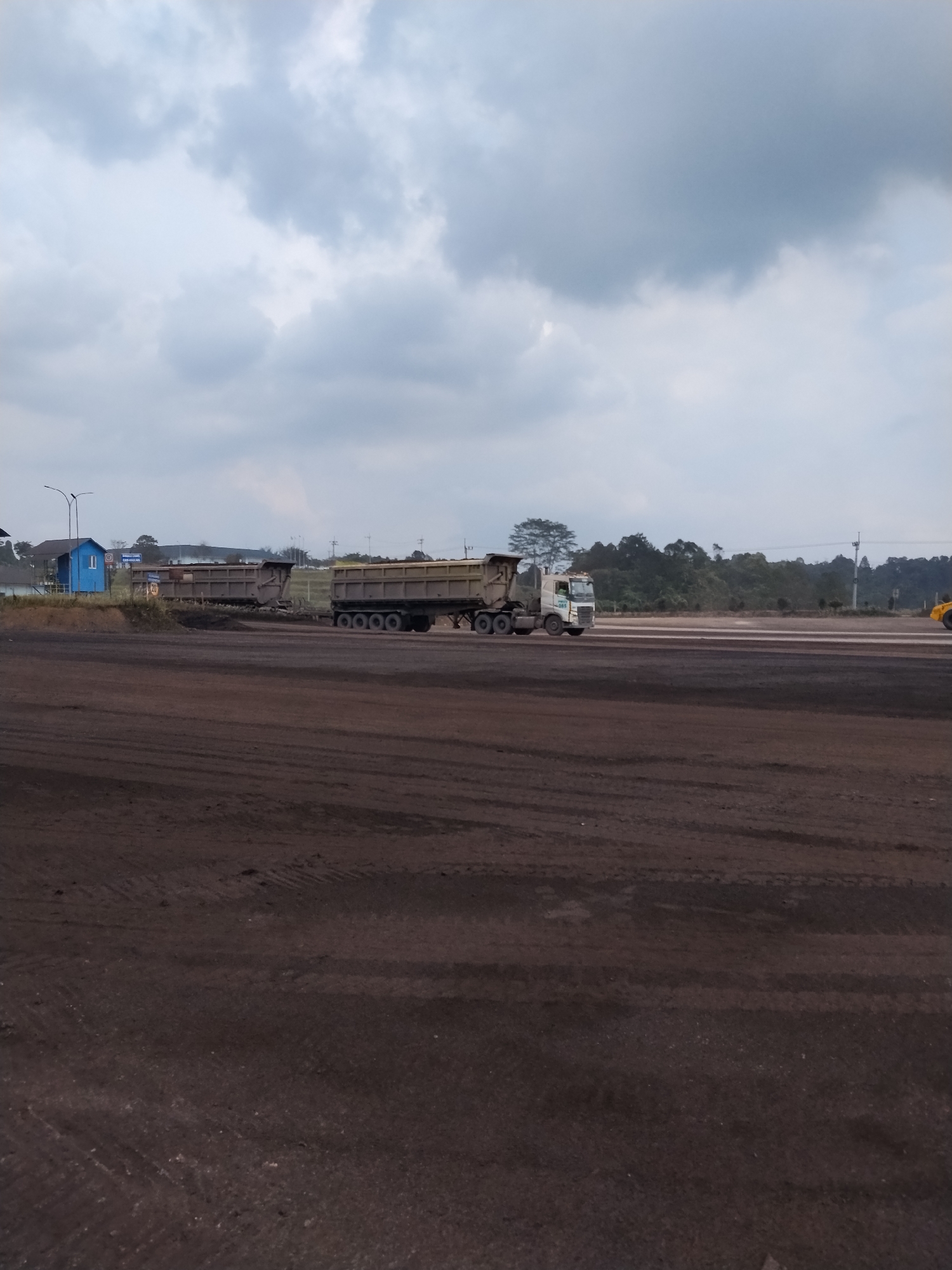 In Sempayau, Indonesia, a wide, flat, dirt expanse stretches across the foreground, marked with tire tracks from heavy machinery. In the mid-ground, two large dump trucks are visible. The truck on the left is partially obscured by the truck on the right, which is a light-colored vehicle with a tan trailer. The truck on the right has its bed tilted upward, suggesting it might be in the process of unloading or has recently finished unloading. Behind the trucks, a line of trees forms the horizon, with a few buildings and industrial structures visible in the distance on the left side of the frame. The sky is overcast with various shades of gray clouds, indicating a potentially cloudy or impending weather event. The overall scene depicts an industrial or mining area, likely at an operational site.