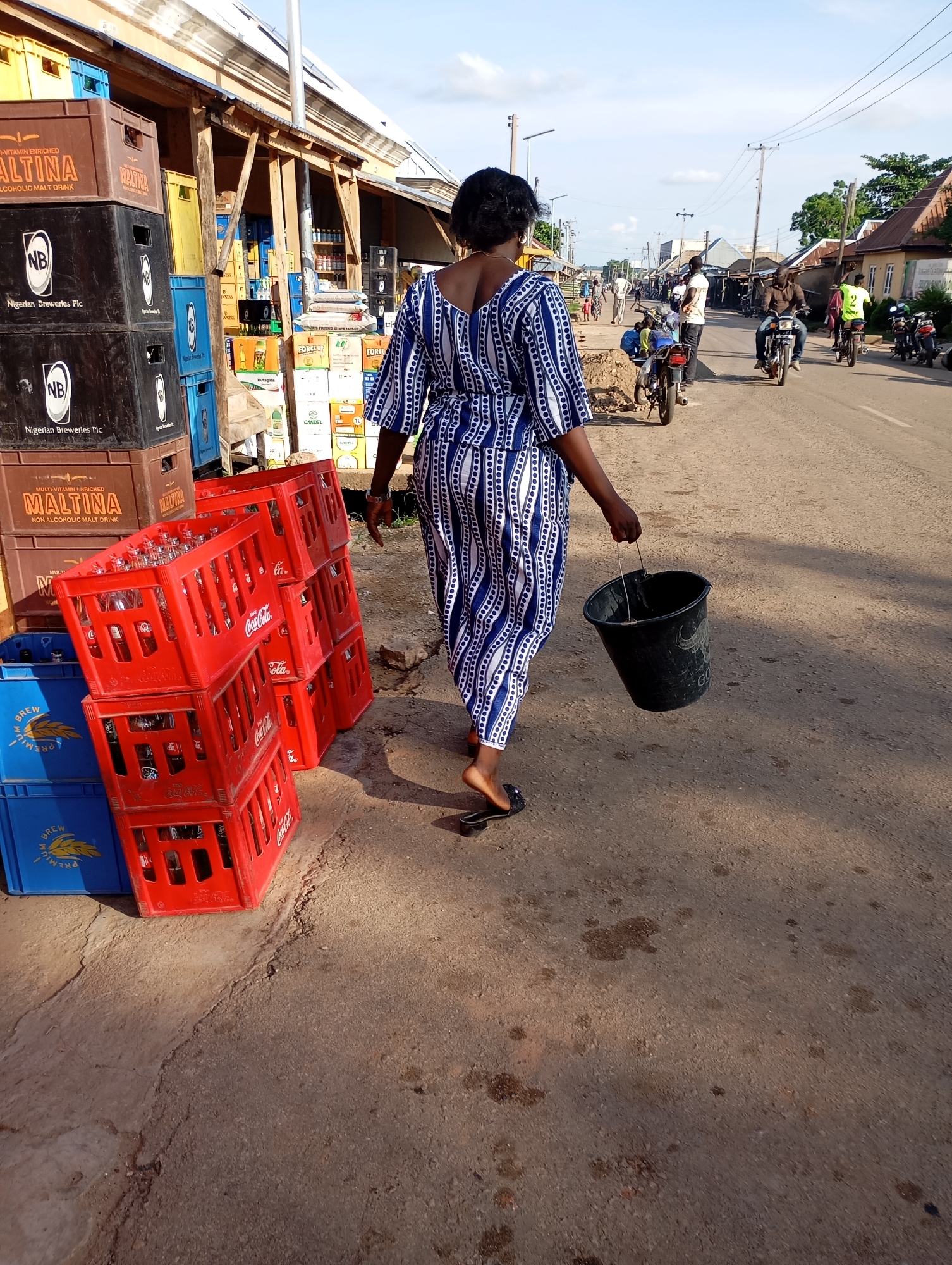 The image depicts a street scene in Shendam, Nigeria, with a woman walking away from the camera, carrying a black bucket. She is wearing a blue and white striped dress and flip-flops. To her left, stacks of plastic crates are visible, some containing bottles. In the background, other people and motorcycles are present on a paved road lined with buildings and shops. The sky is partly cloudy.