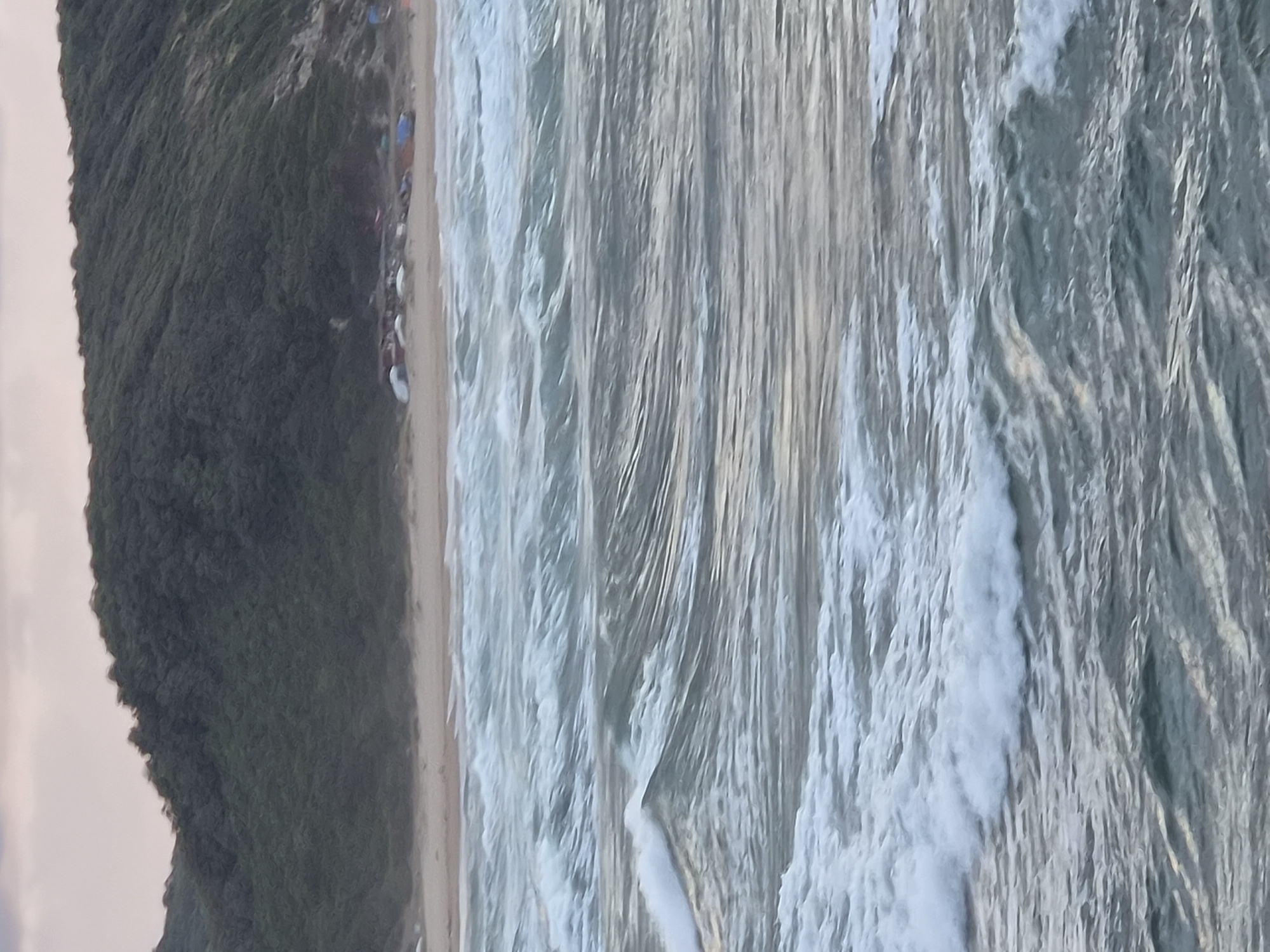 The image depicts a wide shot of the Black Sea coast in Şile, Turkey, during what appears to be dusk. The foreground is dominated by the choppy, grey-green water of the sea, with multiple lines of waves creating a textured surface. The waves are breaking gently, with white foam visible on some of the crests. In the mid-ground, a sandy beach stretches along the coastline, backed by a dense, dark green, tree-covered hill that rises steeply. A cluster of small, colorful tents and what appear to be beach shelters are scattered along the edge of the beach, nestled at the base of the hill. The sky above is a muted pink and grey, suggesting the sun is either setting or has recently set. The overall impression is one of a natural coastal landscape with signs of human habitation in the form of camping.