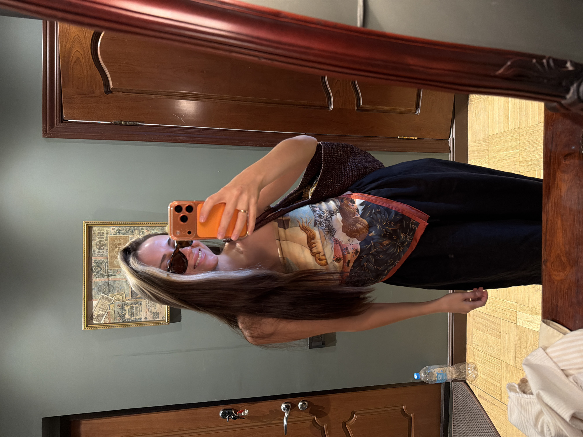 The image shows a young woman taking a selfie in a room. She is wearing sunglasses, a patterned bandana-style top, and a black skirt. She is also carrying a woven bag and holding an orange phone. In the background, there is a wooden door, a framed picture on the wall, and a bottle of water on the floor. The room appears to be a hotel room or an apartment. The floor is made of light-colored wood tiles. The overall lighting is dim. There are no explicit indicators of the location being in Khimki, Russia, within the image itself.