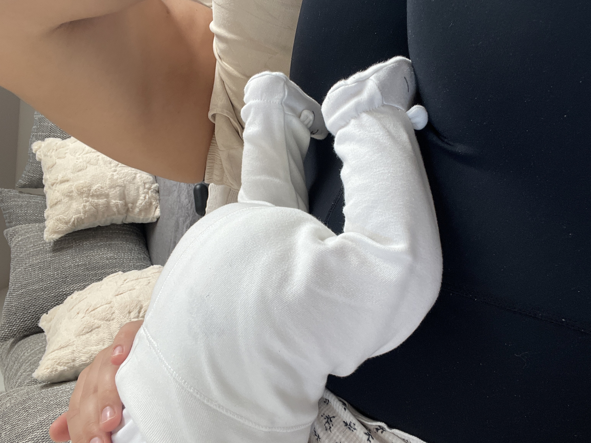 The image shows a close-up of a baby's legs wearing white footed pajamas. The baby's legs are bent at the knees, and the feet are covered by the pajamas. A person's hand is visible in the upper left corner of the image, resting on the baby's leg. The background consists of a couch with several pillows. The scene is indoors and appears to be a home setting. The image does not contain any information about the city of Garmisch-Partenkirchen, Germany.