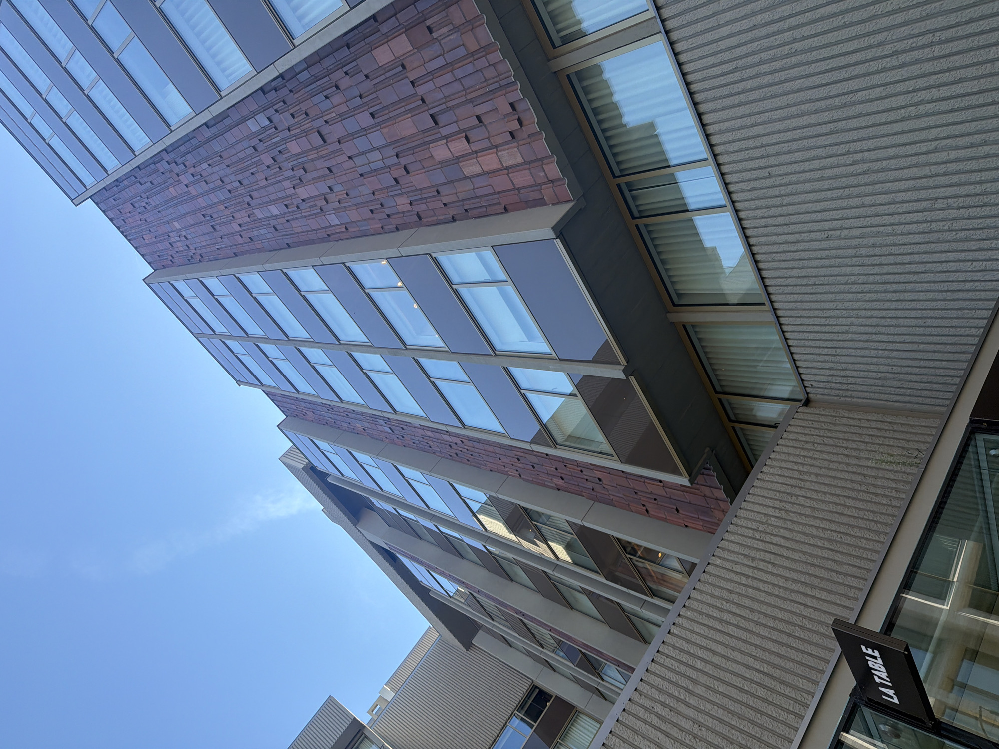 The image shows a low-angle shot of a modern building exterior, likely in Prague, Czechia, under a clear blue sky. The building features a multi-story facade with alternating sections of dark gray siding with large windows and a textured reddish-brown material with a pattern of protruding rectangular blocks. The windows appear to be floor-to-ceiling, with white curtains visible behind them. In the lower left corner, a black sign with white lettering reads "LA TABLE," suggesting the presence of a restaurant or establishment on the ground floor. The building is constructed with a combination of concrete and glass. The overall aesthetic is contemporary and urban.