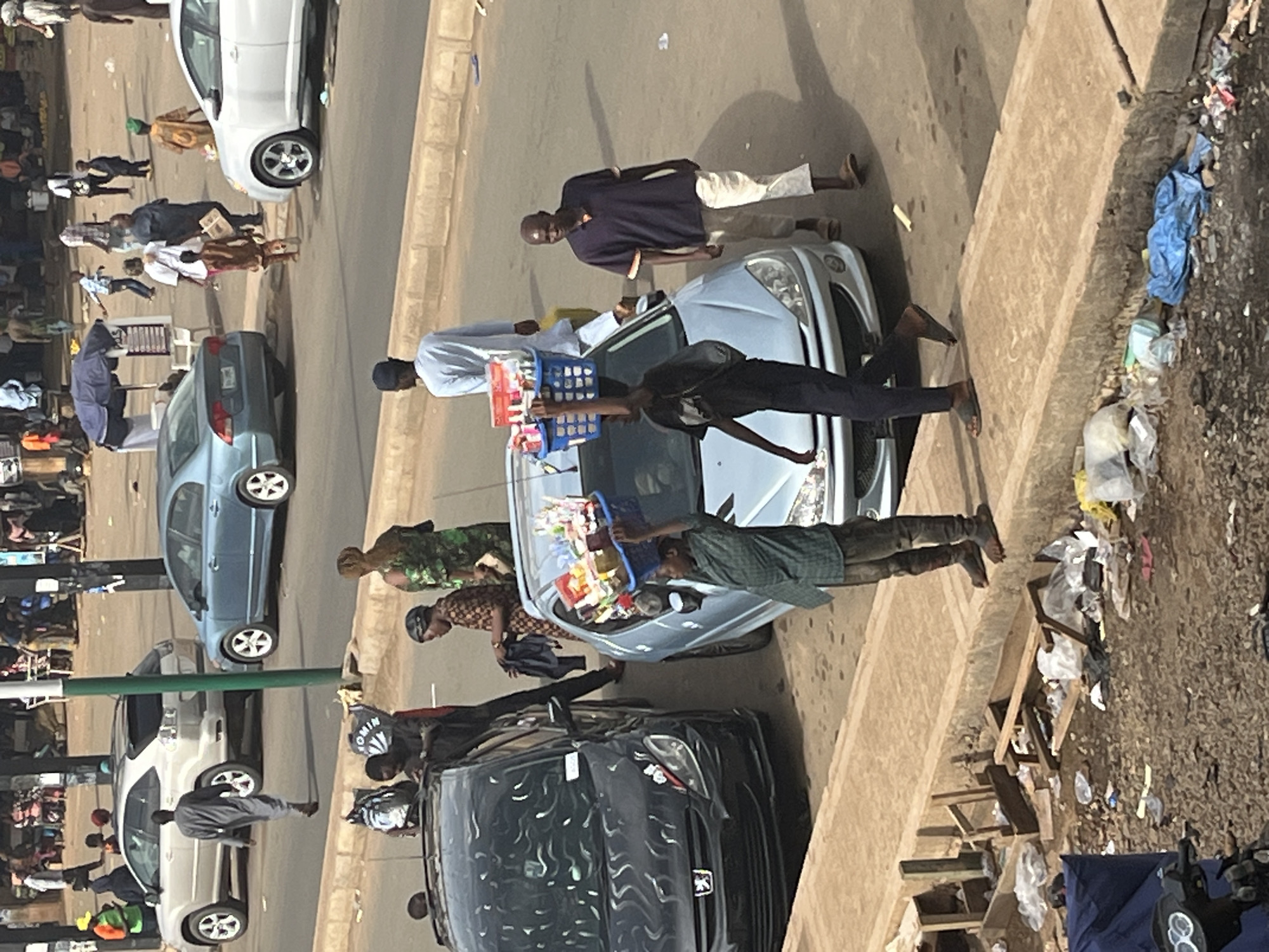 The image captures a street scene in Jos, Nigeria, with a focus on pedestrian and vehicular activity. In the foreground, a young person walks towards the left, balancing a blue basket filled with various items, likely goods for sale, on their head. They are walking on a sidewalk next to a light blue car. To the right, a man wearing a dark blue polo shirt and light-colored pants walks to the left, looking towards the viewer. Behind them, a black car is parked. Several other individuals are visible in the background, including a woman in a green dress and a man in a light blue robe. The street is paved, and there are parked cars, suggesting an urban environment. The ground on the sidewalk area is littered with debris, including plastic bags and wooden scraps. The overall atmosphere appears to be that of a bustling market or commercial area.