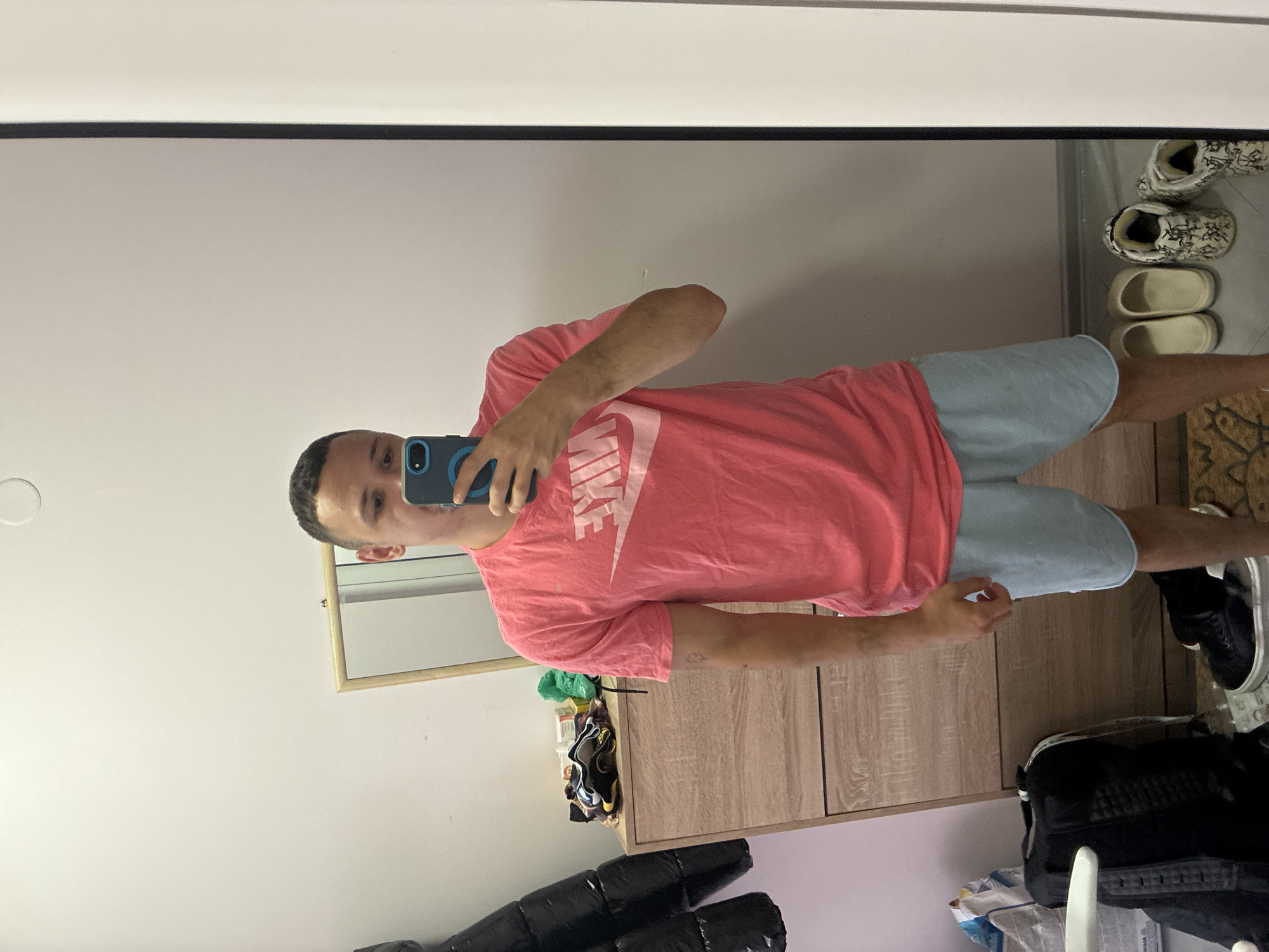 The image displays a full-body mirror selfie of a person standing in an indoor setting, likely in Blagoevgrad, Bulgaria. The individual, who appears to be male, is wearing a red/coral short-sleeved t-shirt with the word "NIKE" prominently displayed in white text across the chest, and light blue shorts. They are holding a blue smartphone with a visible black camera module in front of their face, partially obscuring it, as they capture their reflection.
To the right side of the frame, a light wood-grained dresser or cabinet is visible. On its surface or the floor nearby, several pairs of shoes are arranged, including light-colored slip-on clogs and white and black patterned boots. The person's right foot, clad in a black and white sneaker, is also visible. In the lower left, a black puffer jacket or coat, along with some plastic bags, are stacked on what appears to be the floor or another piece of furniture. A smaller rectangular mirror with a wooden frame is mounted on the wall behind the person's upper body, reflecting a portion of the white ceiling. A patterned mat or rug is partially visible on the floor to the right. The scene depicts a casual interior, likely a bedroom or dressing area. The time of day is not discernible due to indoor lighting.