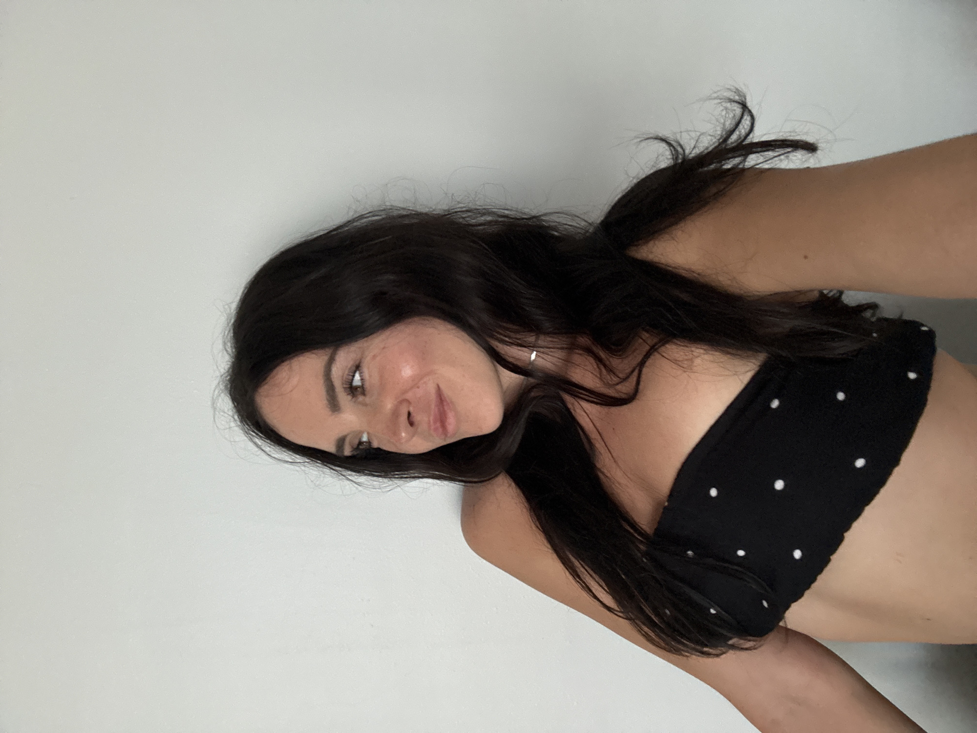 In North Kuta, Indonesia, a young woman with long, dark hair is captured in a mid-shot against a plain, light-colored wall. She is wearing a black bandeau top with white polka dots. Her body is angled to her left, while her head is turned to her left, with her gaze directed slightly upwards. Her left arm is bent, with her hand resting behind her head, and her right arm is visible on the left side of the frame. A delicate silver necklace is visible around her neck. The lighting suggests an indoor setting, possibly a home or studio, with natural light illuminating her face.