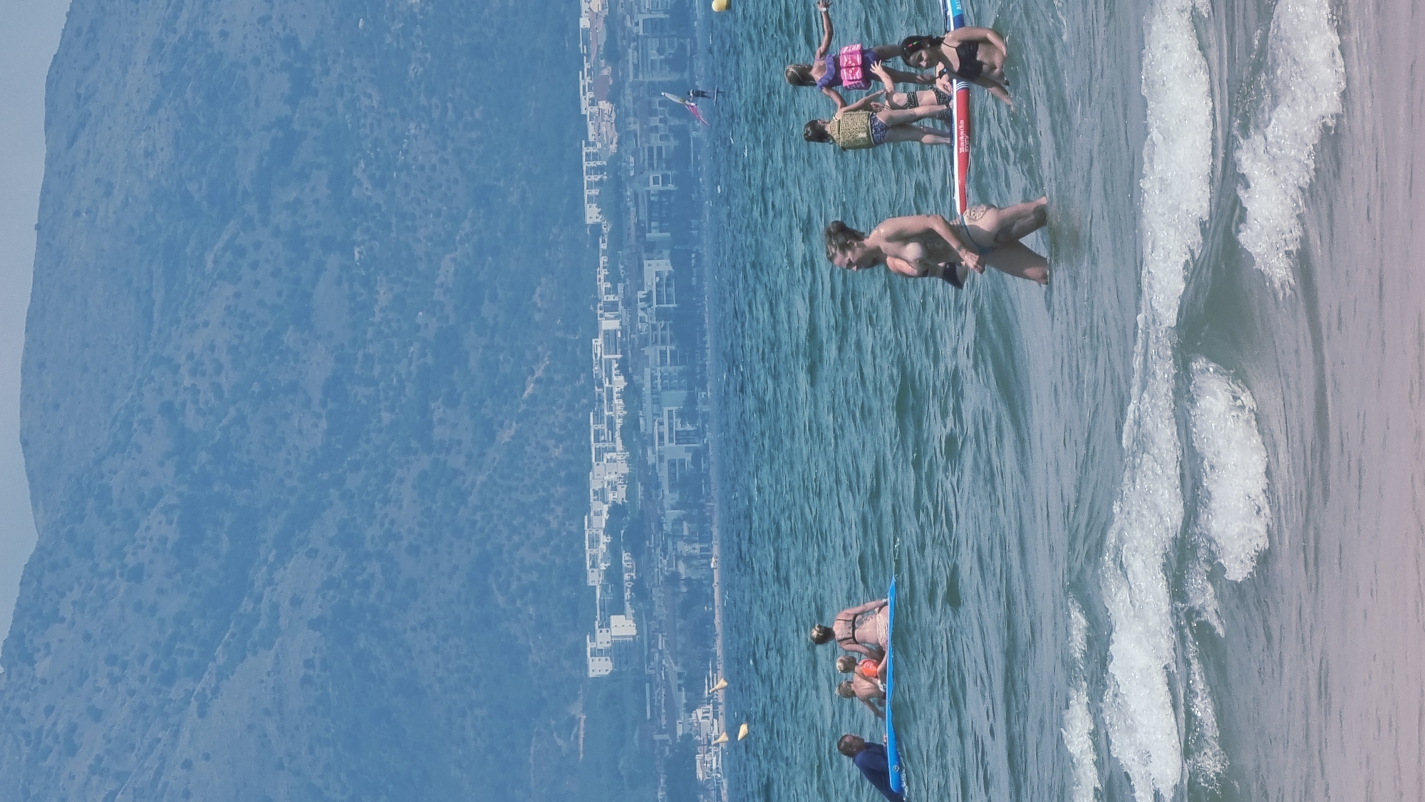 The image captures a scene at the beach in Castelló d'Empúries, Spain, with a mountainous backdrop. In the foreground, the ocean is visible with moderate waves. Several people are in the water. On the left, a group of four is on a blue inflatable. Three are children and one adult male is partially visible on the left. Further right, a woman is standing in the water. To her right, two young girls are on a paddleboard, with one appearing to be an older child or teen. Another woman is partially visible behind them. In the mid-ground, a windsurfer is visible on the water, and yellow buoys are scattered across the sea. In the background, a coastal city with white buildings is situated at the base of the mountains. The sky is clear and bright.