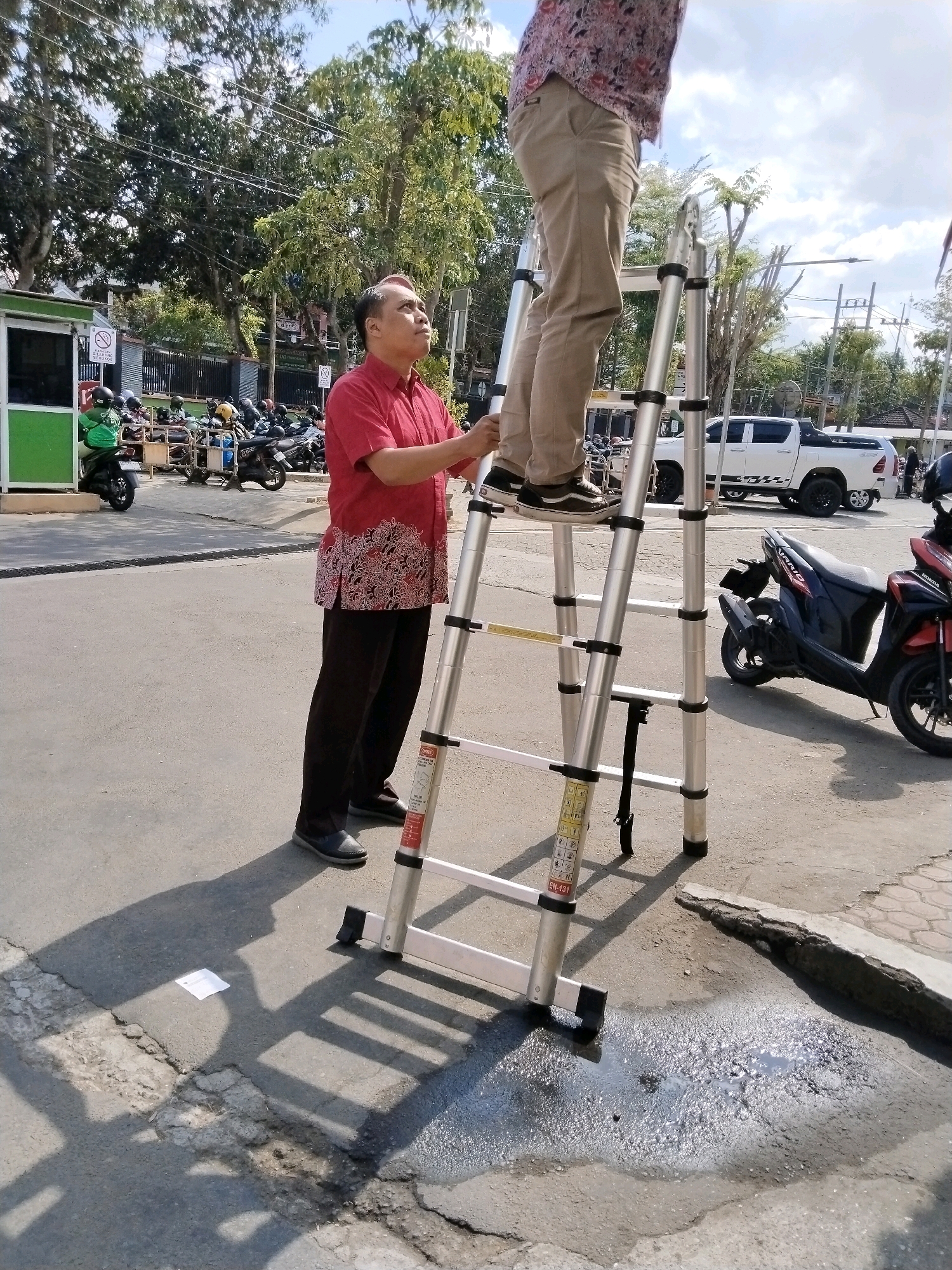 The image depicts an outdoor scene in Trenggalek, Indonesia, likely on a street or public area, given the presence of parked motorcycles and a white pickup truck in the background. Two men are the central figures. One man, wearing a red short-sleeved shirt with a white and red patterned hem and dark trousers, stands on the ground next to a tall, silver, extendable ladder. He is looking upwards. The second man, wearing a patterned shirt and beige trousers, is standing on the ladder, also looking upwards. The ladder is a significant object, angled and appearing to be in use or ready for use. The environment suggests a sunny day with some trees and utility poles visible. A green structure, possibly a kiosk or guardhouse, is located on the left side of the frame, with motorcycles parked nearby. There are signs on the green structure and on the ladder itself. The ground is asphalt with some cracks and a puddle of water. The scene appears to be of everyday life in an Indonesian urban or semi-urban setting.