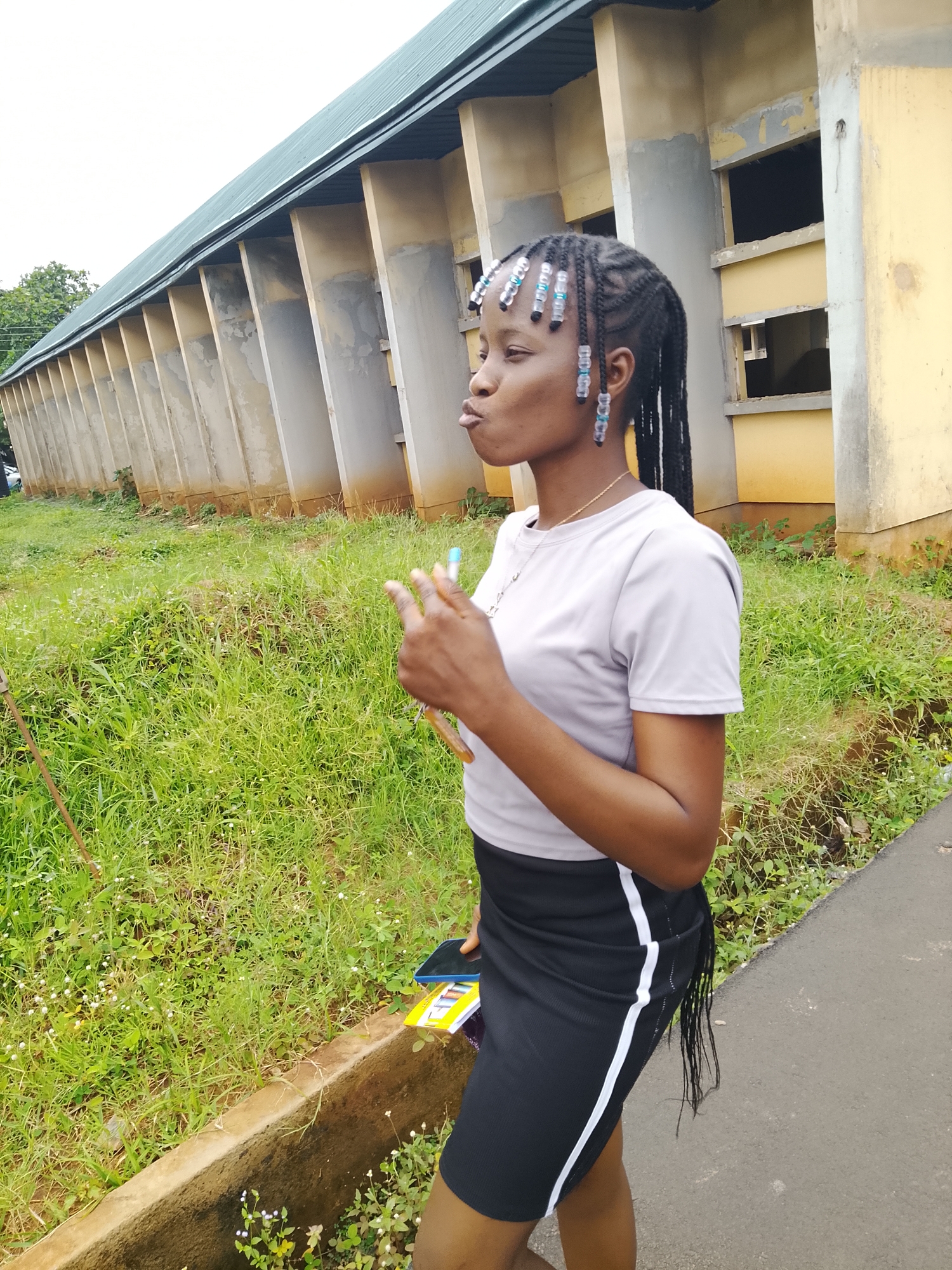 A medium shot captures a young Black woman from the side, standing on a paved surface next to a grassy area. She has her hair in braids adorned with blue and clear beads, and she is wearing a light gray t-shirt and a black skirt with white stripes. Her mouth is slightly open, as if she is speaking or making a gesture. She is holding two pens or markers in her right hand, and a cell phone and a notebook or folder are tucked under her left arm. Behind her, a light yellow building with a green roof runs along the length of the frame. The building has a repetitive pattern of concrete pillars and open window frames, suggesting it may be an academic institution or an abandoned structure. The grassy area in front of the building is lush and overgrown, with scattered small white flowers. A concrete curb separates the paved area from the grassy section. The overall impression is one of an outdoor setting in a somewhat neglected environment.