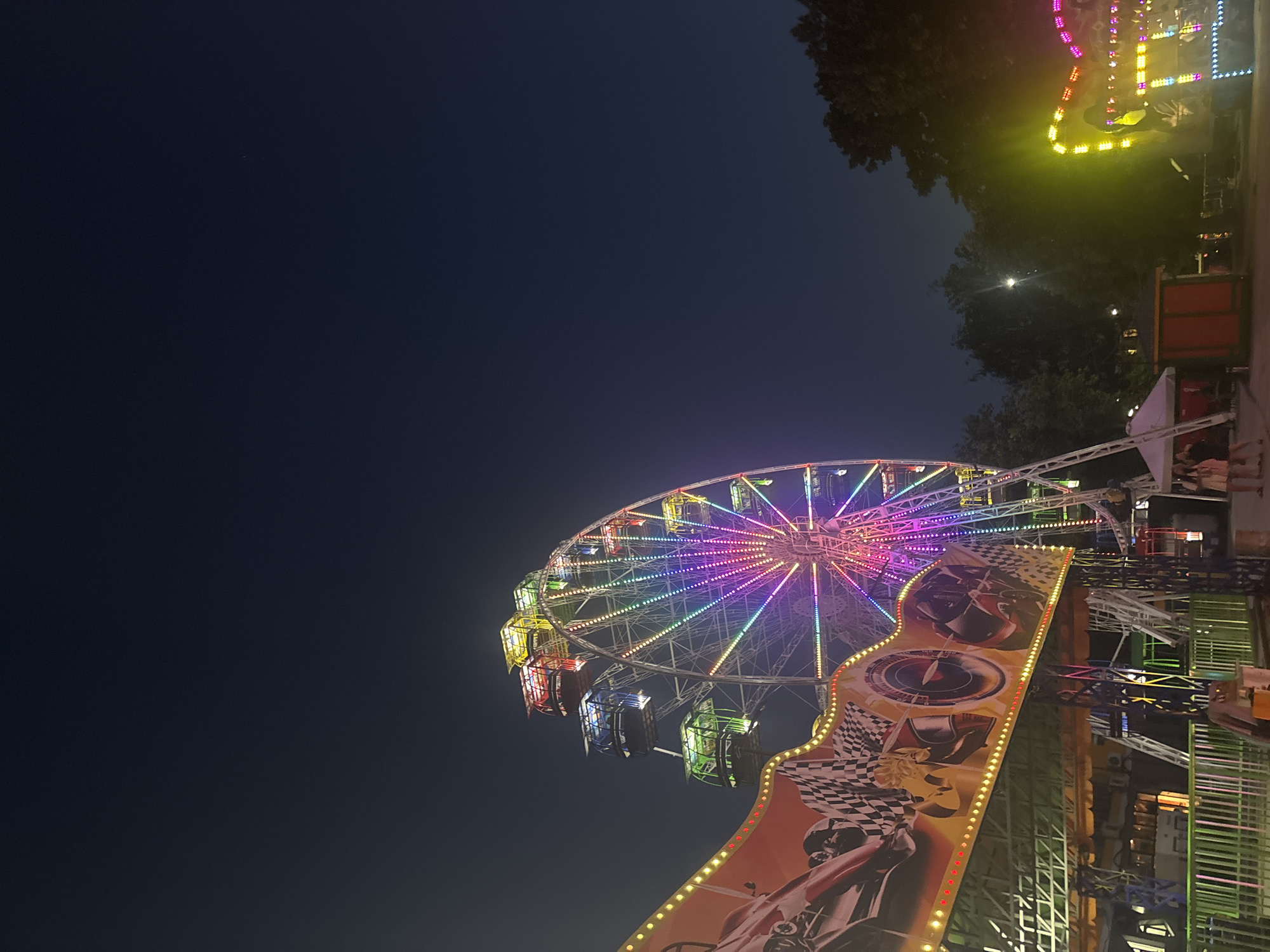 A ferris wheel, illuminated with colorful lights, dominates the scene at a nighttime amusement park. The ferris wheel is situated in the foreground, with its bright lights contrasting against the dark night sky. In front of the ferris wheel, a banner advertising a racing theme is displayed, featuring images of cars and checkered flags, also lit with colorful lights. To the right of the ferris wheel, more amusement park rides are visible, also adorned with bright lights. The overall environment suggests a lively and festive atmosphere, likely a popular attraction in Bila Tserkva, Ukraine. The image captures the essence of a fun-filled evening at an amusement park, with the ferris wheel serving as the central focal point.