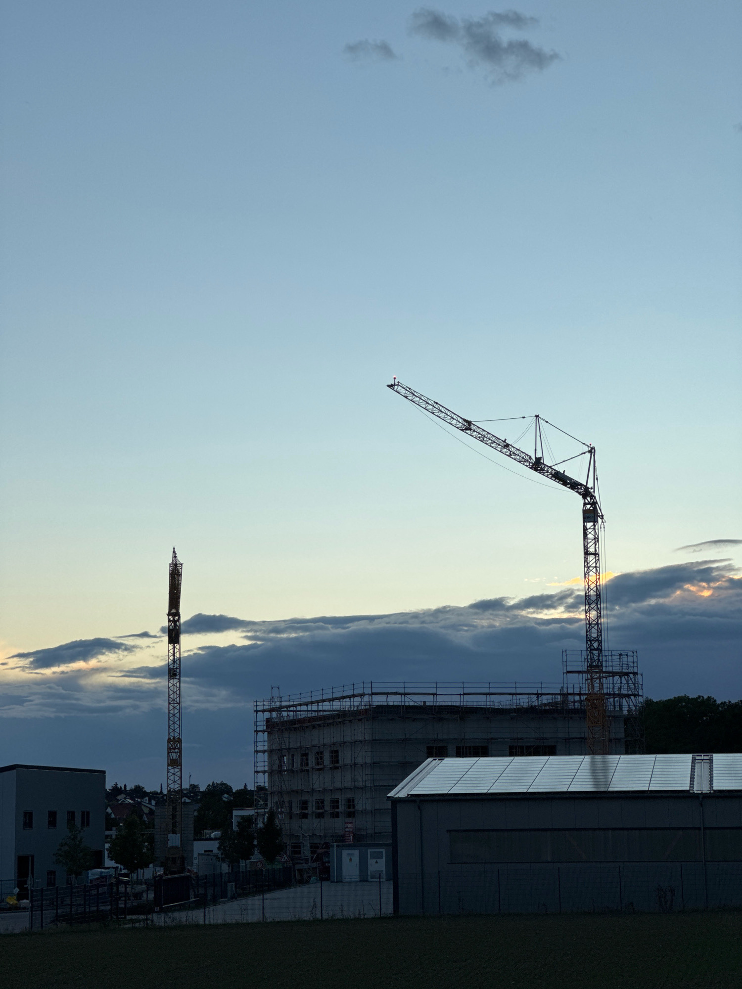 The image depicts an outdoor construction site in Lenting, Germany, at dusk. In the foreground, a dark, grassy field is visible, with a chain-link fence running along its edge. Behind the fence are several low-rise buildings. One building on the right has a dark gray, angular roof covered with solar panels, and a smaller white structure attached to its side. To the left of this building is a larger, gray building with a fence in front of it.
Dominating the middle ground are two construction cranes. The taller, thinner crane, a lattice tower, stands vertically on the left side of the frame, with its top section silhouetted against the sky. To its right, a larger, more complex construction crane with a long jib extends diagonally across the frame, its boom angled upwards and to the left. Both cranes are positioned near a multi-story building under construction, which is partially obscured by scaffolding. This building appears to have a concrete facade with visible window openings.
The sky above is a gradient of pale blue near the horizon, transitioning to a darker blue towards the top. Scattered clouds are present, with some darker, heavier clouds forming a band across the middle of the sky. The setting sun is partially visible behind these clouds, casting a warm, orange glow that is reflected on the clouds and the arm of the larger crane. The overall lighting suggests the scene is captured during the twilight hours. No people are visible in the image.