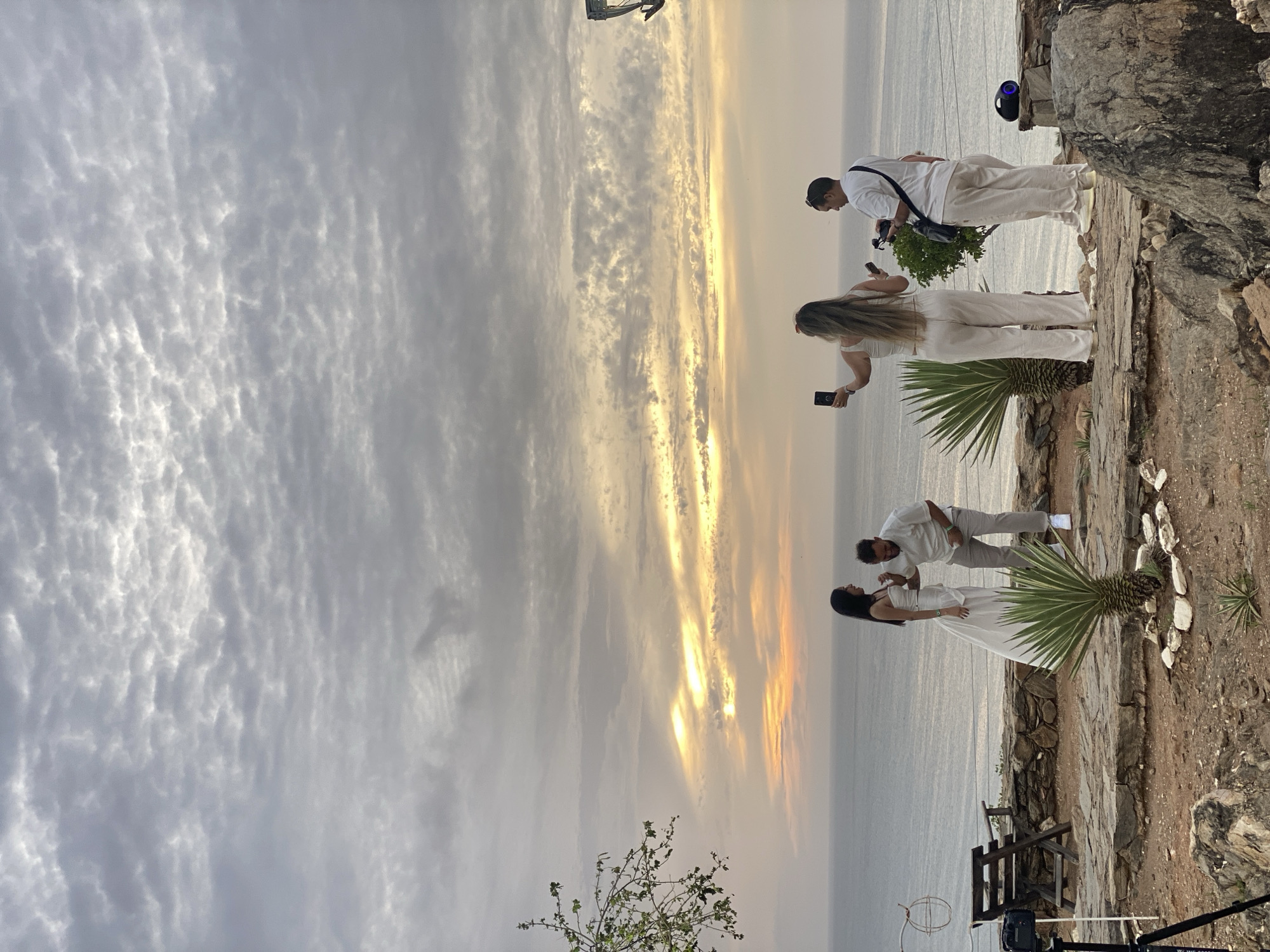 The image presents an elevated coastal scene in Juan Griego, Venezuela, during either sunset or sunrise, characterized by a heavily clouded sky overhead transitioning to warm yellow, orange, and pink light streaks along the horizon above a calm, grey-blue sea.
On a terraced platform constructed from light-colored stone paving and natural rock, four individuals are positioned. On the far left, a black camera is mounted on a black tripod, directed towards the central figures. Behind the tripod, a dark wooden bench and table are visible, with a rusted, circular metal structure partially obscured.
In the center-left, a woman with long dark hair, dressed in a white strapless top and a flowing white skirt, stands facing a man. This man, with short dark hair and a beard, wears a light-colored collared shirt and trousers, leaning towards the woman with his hand on her arm, engaged in an expressive interaction.
To the right of the central pair, a second woman with long, light-colored hair, wearing a white sleeveless top and matching wide-leg pants, holds a black smartphone aloft with both hands, seemingly capturing a photograph or video of the interaction. Further to the right, a second man, wearing a white t-shirt, light-colored pants, and sunglasses on his head, carries a black bag over his shoulder and holds a black electronic device, observing the activity.
The ground consists of reddish-brown soil and large grey rocks, with scattered small, spiky succulents. A prominent agave-like plant grows in a small rock-ringed bed in the center foreground. On the far right, a dark, cylindrical portable speaker emitting a blue light rests on a rock formation. A tree branch with green leaves extends into the frame from the left side. The scene suggests a planned photographic or video production, possibly a wedding or engagement shoot.