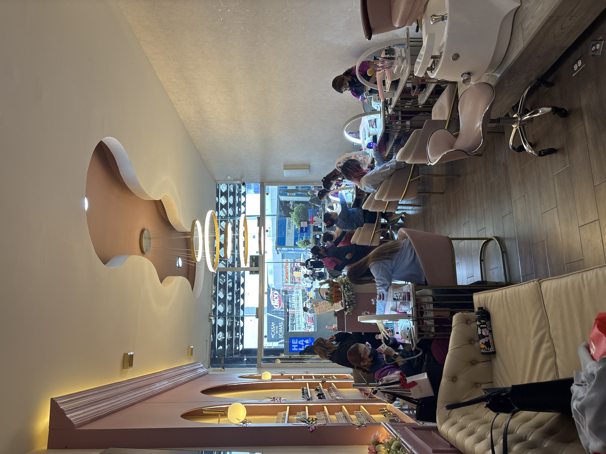 The interior of a nail salon in San Bernardino Tlaxcalancingo, Mexico, shows a row of customers receiving manicures. The salon has large windows revealing a street scene. People are seated at manicure stations, and technicians are attending to their nails. The salon has a pink and beige color scheme. Display shelves with nail polish are visible on one side. Overhead, there are circular light fixtures. A sofa is positioned near the manicure stations.