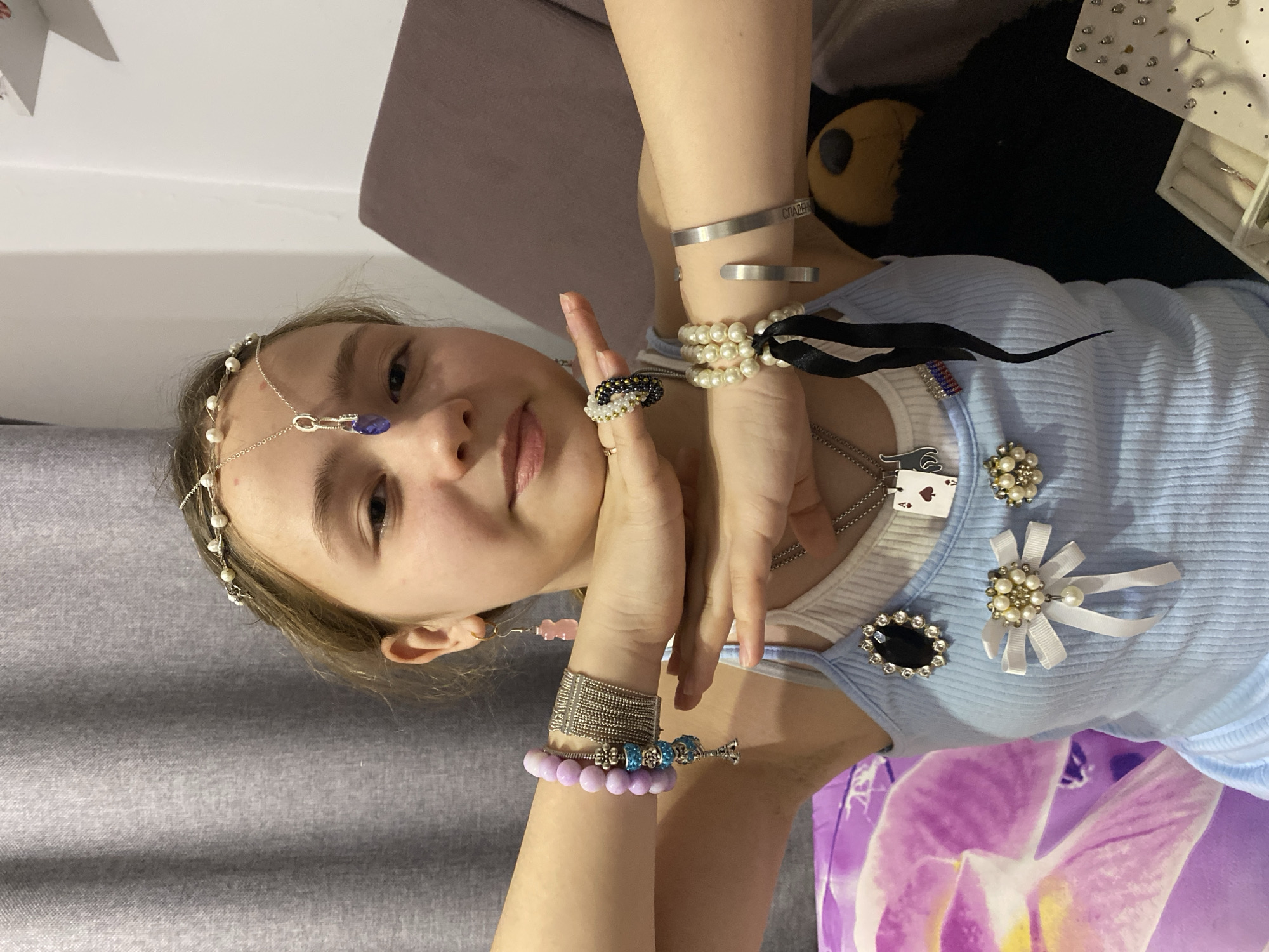 A young girl is shown in a portrait-style photograph. She is wearing a light blue tank top, adorned with various brooches, including one with pearls and white ribbons, and another with a large black gem surrounded by smaller gems. A playing card pendant, shaped like a spade, hangs on a silver chain around her neck.
The girl is wearing numerous pieces of jewelry. On her wrists are several bracelets, including one with light purple beads, another made of silver mesh, and one with blue decorative elements. On her other wrist, she is wearing metal cuffs and a bracelet made of pearls tied with a black ribbon. Rings adorn her fingers, including one with black, gold, and pearl beads. A delicate headpiece with pearls and a blue gemstone is also on her head.
The background is slightly blurred. To the right, part of a black stuffed animal can be seen, as well as some items that look like jewelry on a white display. Behind the girl, there's a gray surface, possibly a wall or piece of furniture. The setting appears to be indoors.