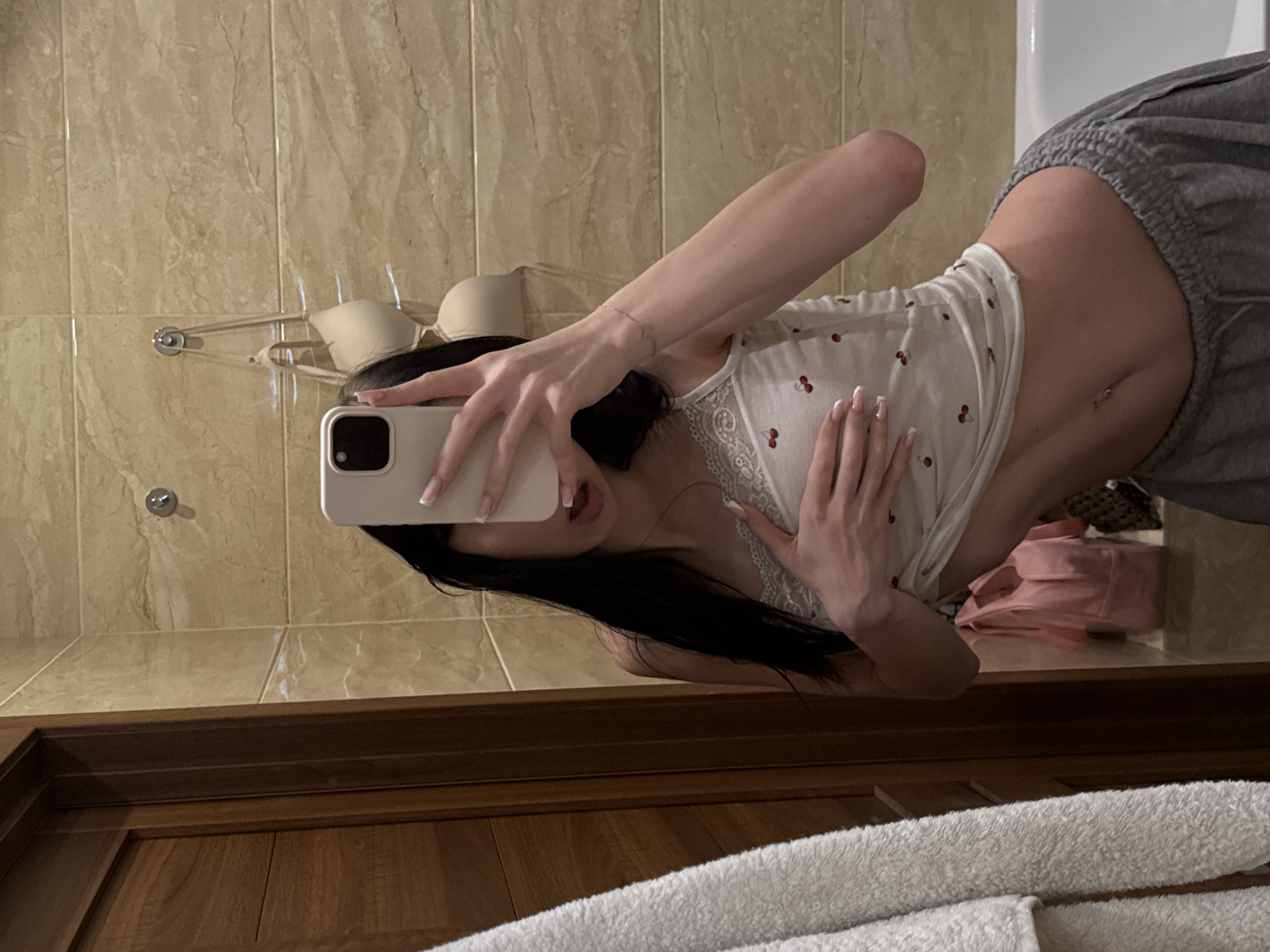 In a bathroom with beige tiled walls, a young woman with long, dark hair takes a mirror selfie. She is wearing a white tank top with a cherry pattern and grey sweatpants. Her left hand is on her chest, and her right hand holds a smartphone in front of her face. A light-colored towel hangs on the left side of the frame, and a wooden door is visible behind it. Two bras hang on hooks on the wall behind her, and a pink bag and a patterned bag are on the floor to her right. A white bathtub is partially visible on the far right. The overall impression is that this is a private moment captured in a residential bathroom.
