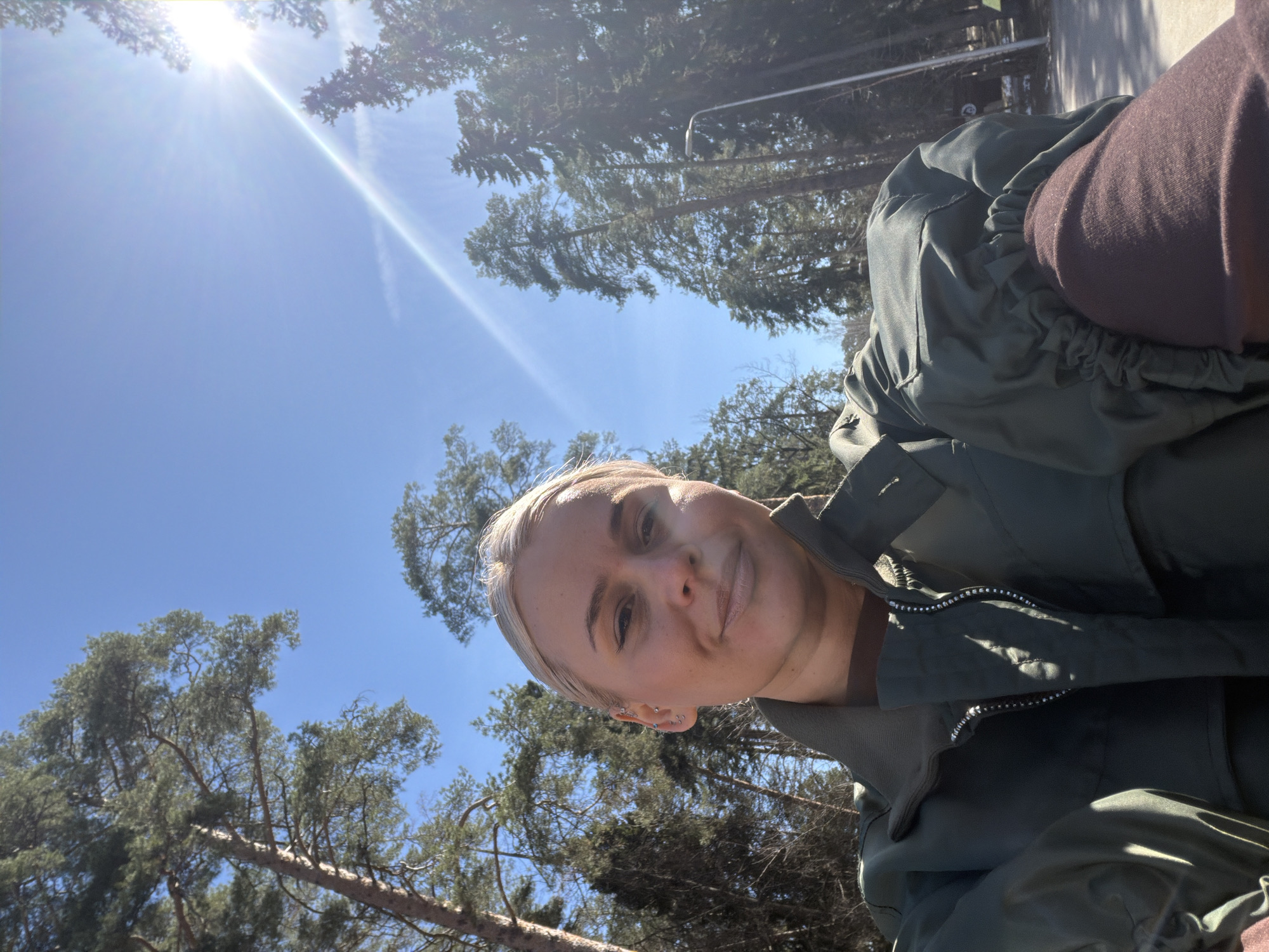 A selfie shows a person with light blonde hair, wearing an olive green jacket and brown turtleneck. The background consists of a clear blue sky and tall trees, some with dense foliage and others bare. The bright sun is visible in the upper left corner. The scene appears to be in a wooded or park-like area. There is insufficient information to determine if this photo was taken in the city of Sarapul, Russia.