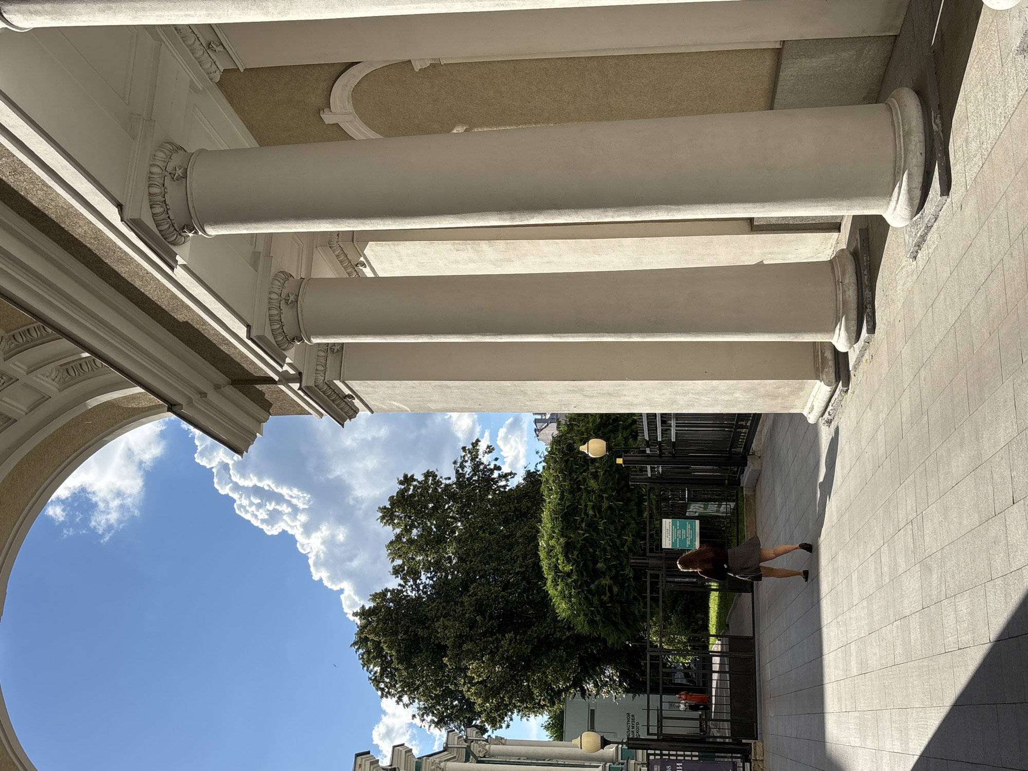 The image shows a pedestrian walkway leading towards a large gate with a woman walking away from the camera. The walkway is paved with light-colored square tiles and is partially covered by shadows. To the right of the walkway are several large, cream-colored columns with ornate capitals, part of a classical-style building. Beyond the gate, there are trees and a building with a sign. The sky above is blue with large, puffy white clouds. The overall scene suggests an outdoor public space, possibly a park or the grounds of a cultural institution, with elements of classical architecture. The location is identified as Novaya Usman', Russia.