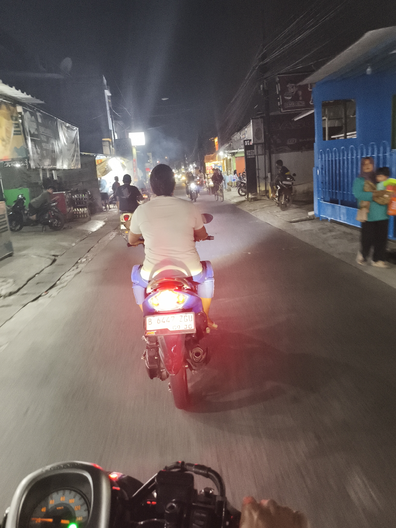 The image depicts a street scene at night in Depok, Indonesia, likely along Jalan Margonda or a similar main thoroughfare. The perspective is from the viewpoint of someone riding on a motorcycle, looking forward and slightly down. The main subject in the foreground is the back of another motorcycle rider wearing a white t-shirt and blue shorts, also on a motorcycle. The license plate of this motorcycle reads "B 6447 ZGU 09-26".
To the left of the road, there are various buildings and structures. Some appear to be shops or stalls with awnings and signage, dimly lit. A few other motorcycles and people are visible further down the street. On the right side of the image, a blue building with a barred entrance stands out. A woman holding a baby or small child is visible near this building.
The road surface is asphalt, with some white markings indicating lanes. Streetlights and lights from the shops illuminate the scene, creating reflections and shadows on the road. The overall atmosphere is that of a busy urban street at night, with moderate traffic and pedestrian activity. The sky is dark, indicating it's nighttime. The image has a slightly grainy quality due to low light conditions.